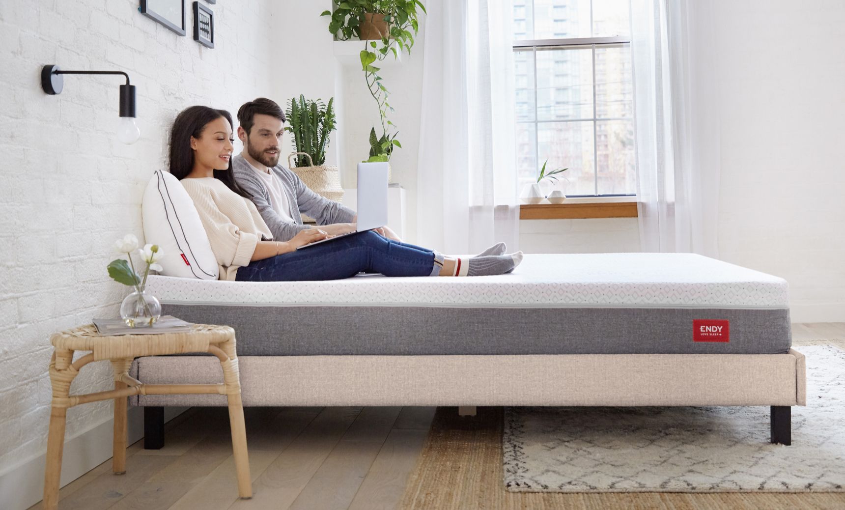 A couple sitting on a bed in a room, browsing on their laptop.