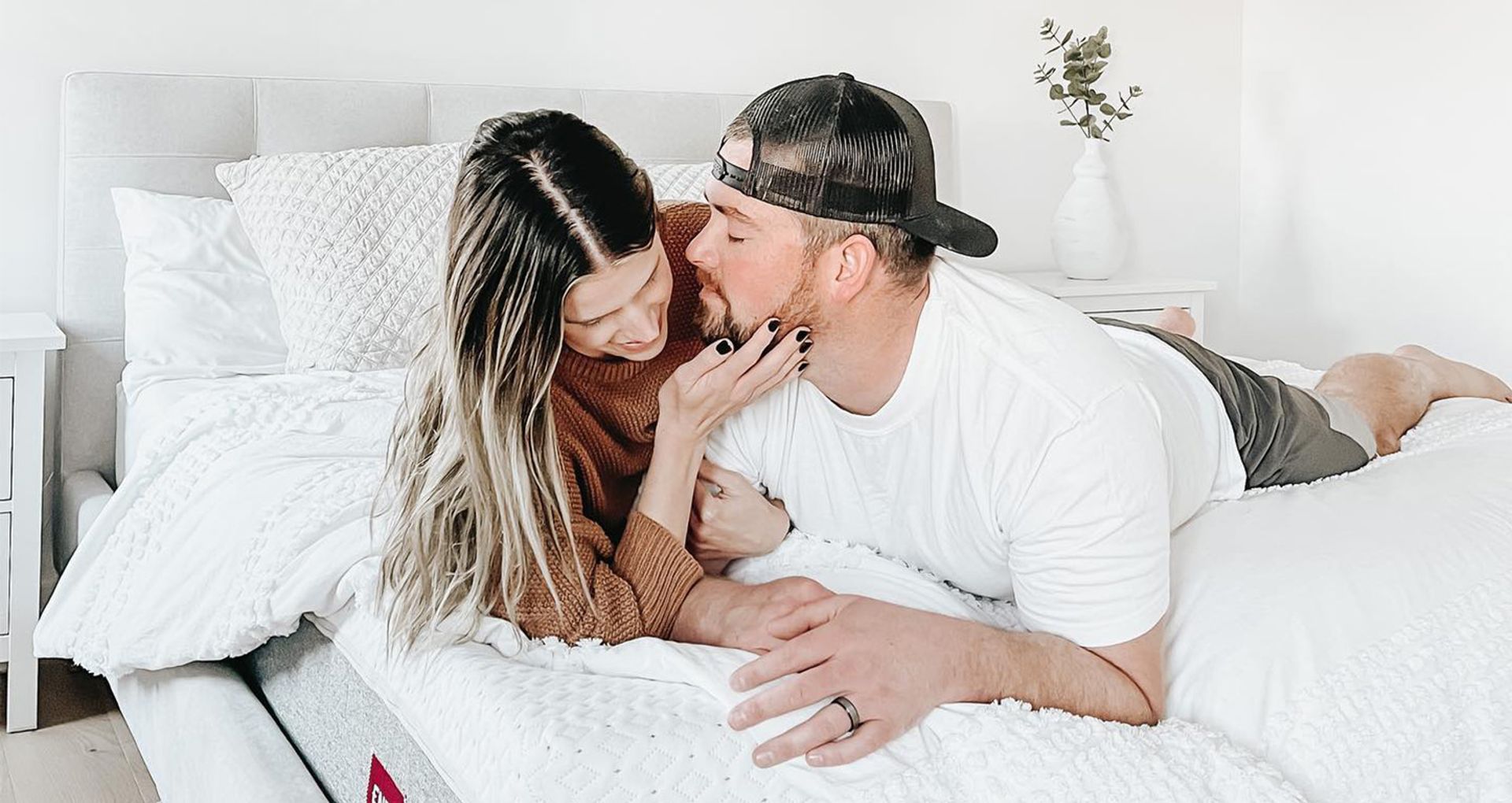  A couple lying on their stomachs in bed, one kissing the other on the cheek.