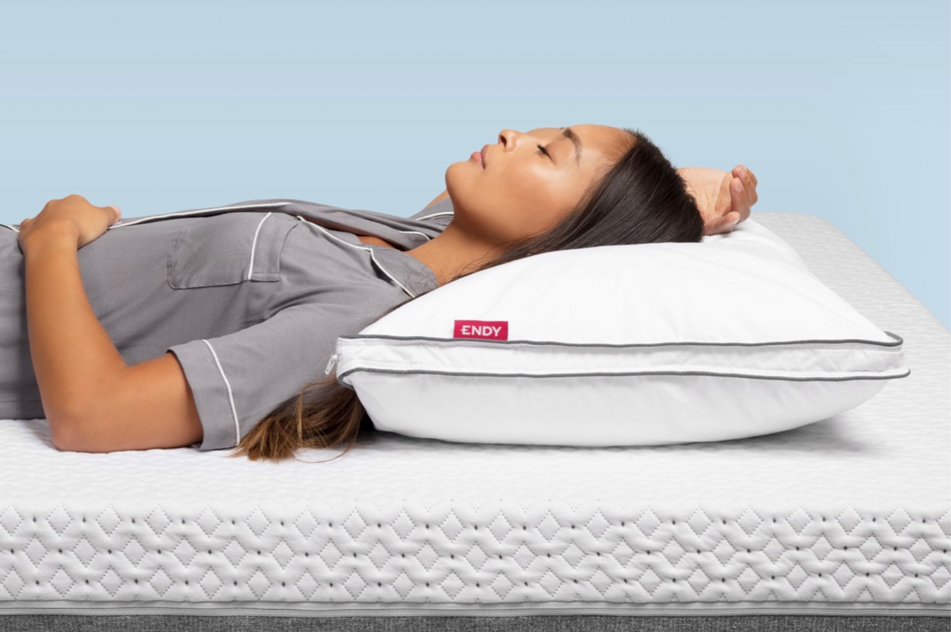 Tan woman with black hair, wearing grey pajamas, lying on an Endy Mattress with an Endy Customizable Pillow under her head