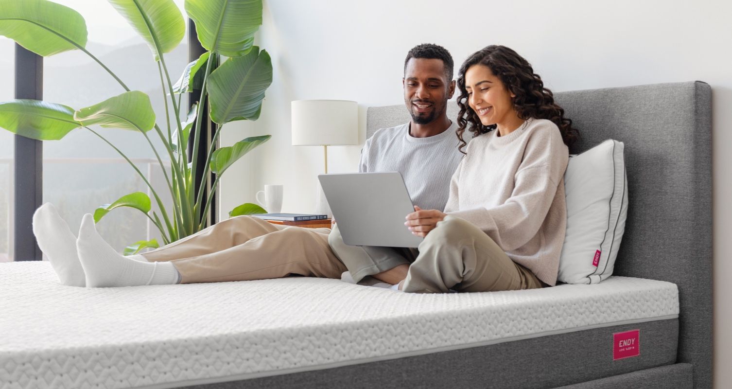A man and woman sitting up in bed, looking at a laptop screen.