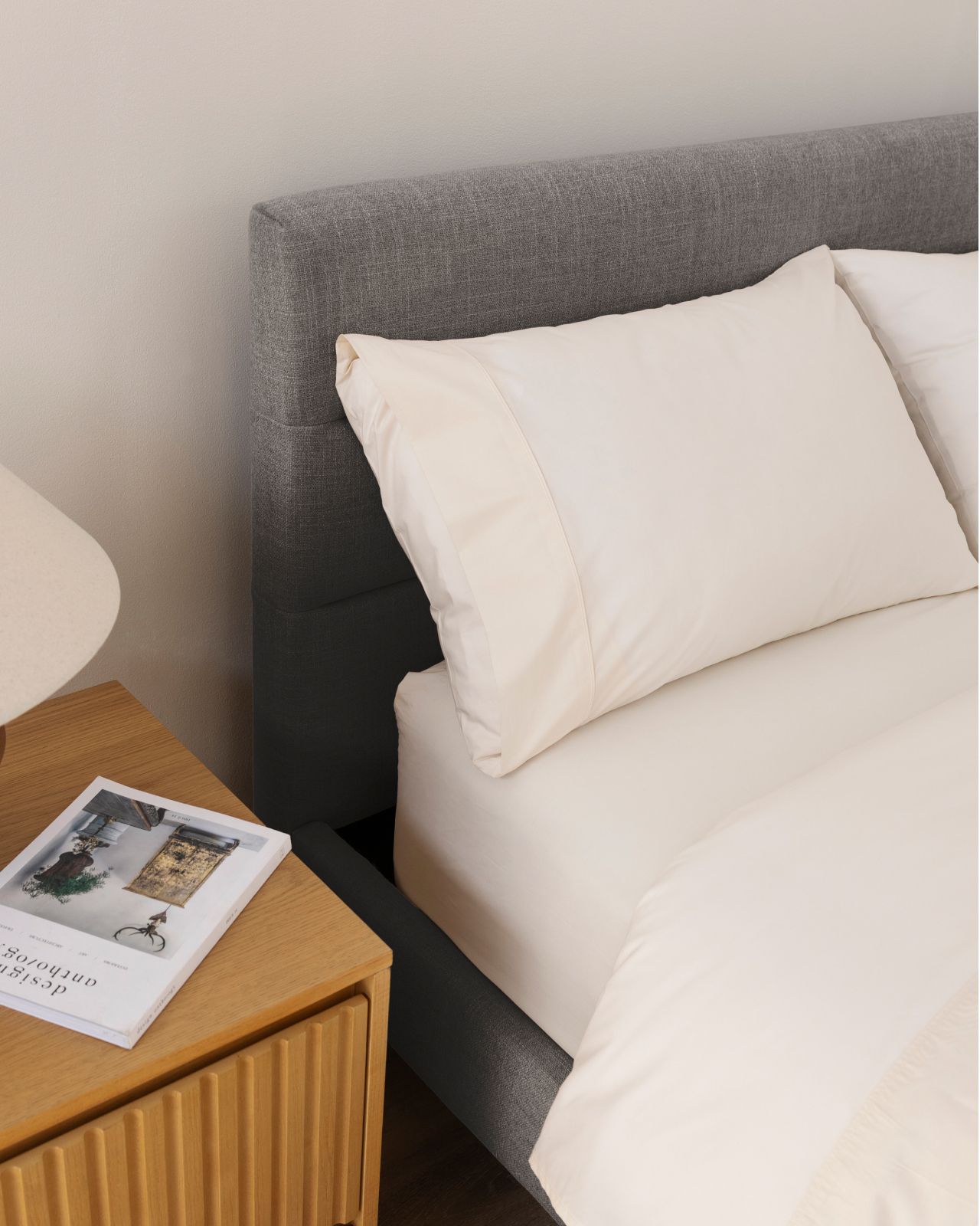 Bedroom with channel upholstered bed frame in graphite colourway, shown with wooden nightstand and soft cream sheets.