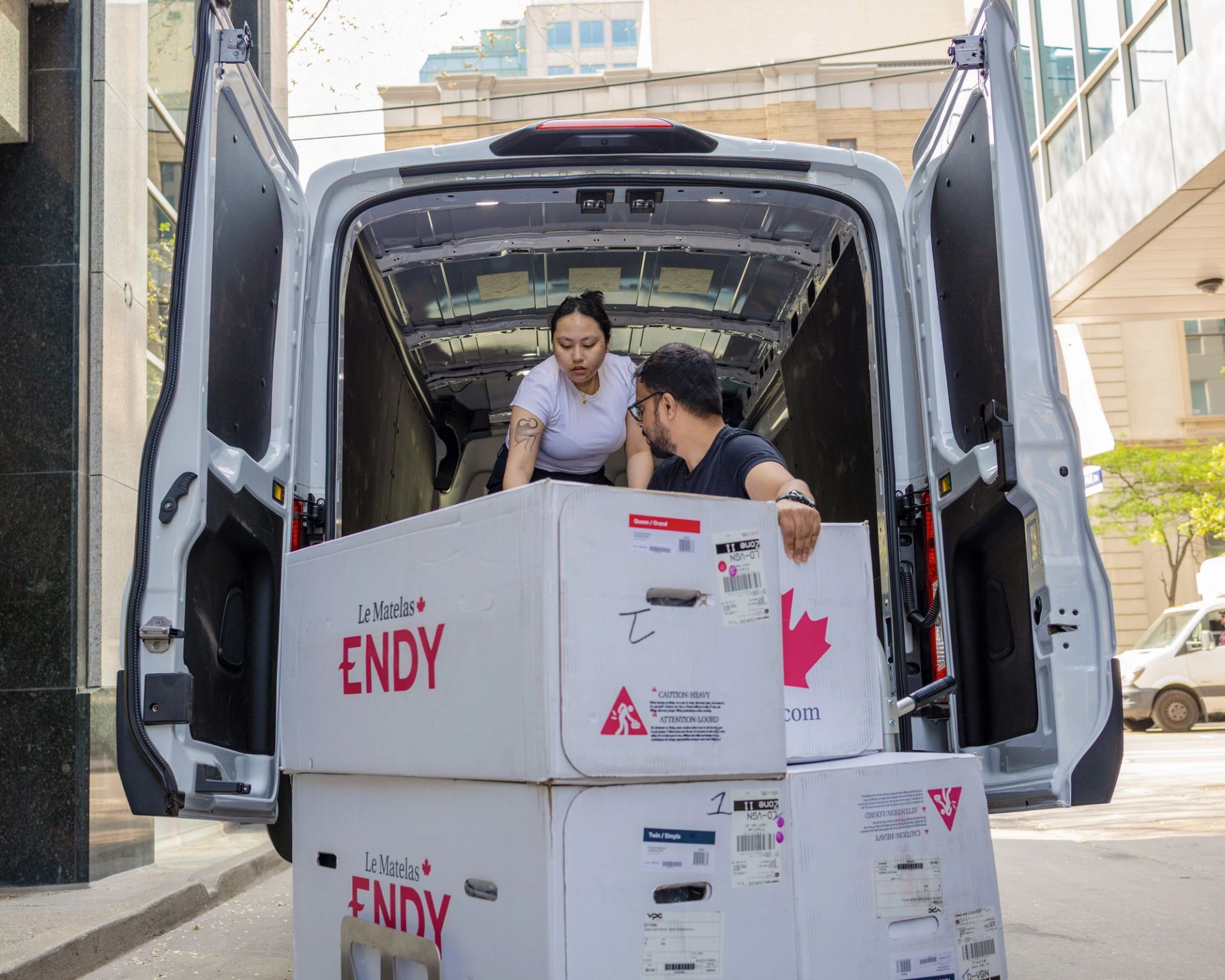 Two people loading four Endy Mattress boxes into the back of a white van.