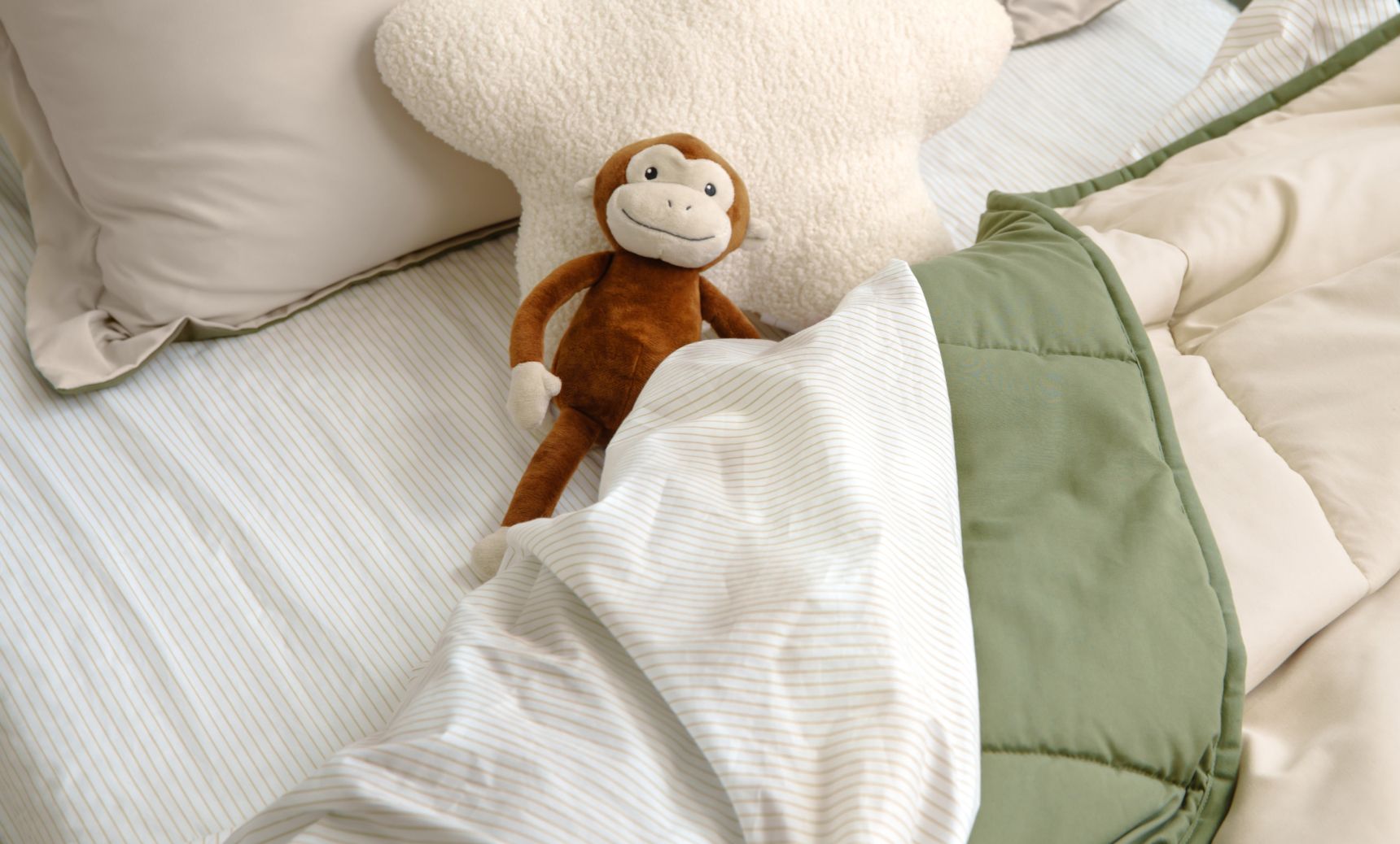 Close up of a children's bed with striped sheets, soft green cover and a monkey stuffed animal on the pillows