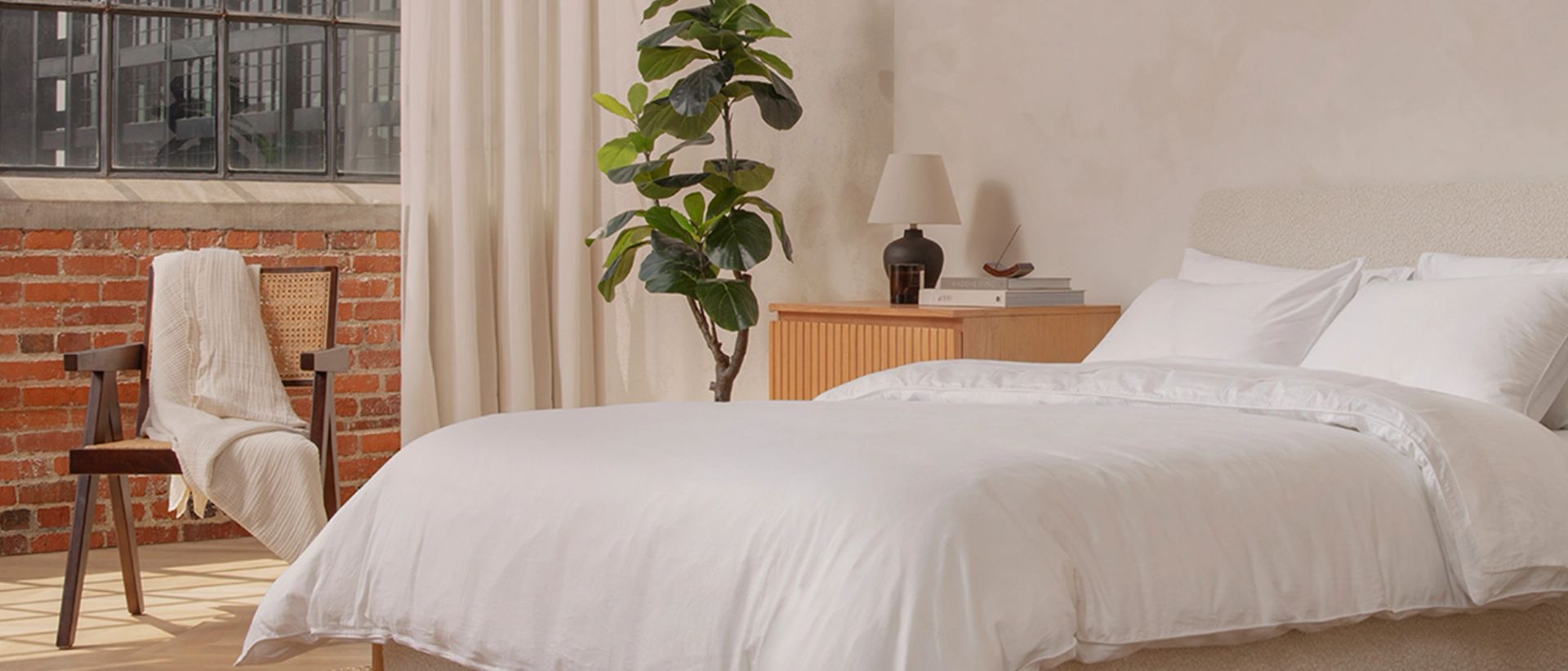 A bed with white sheets, next to a lush plant and chair with blanket draped over it