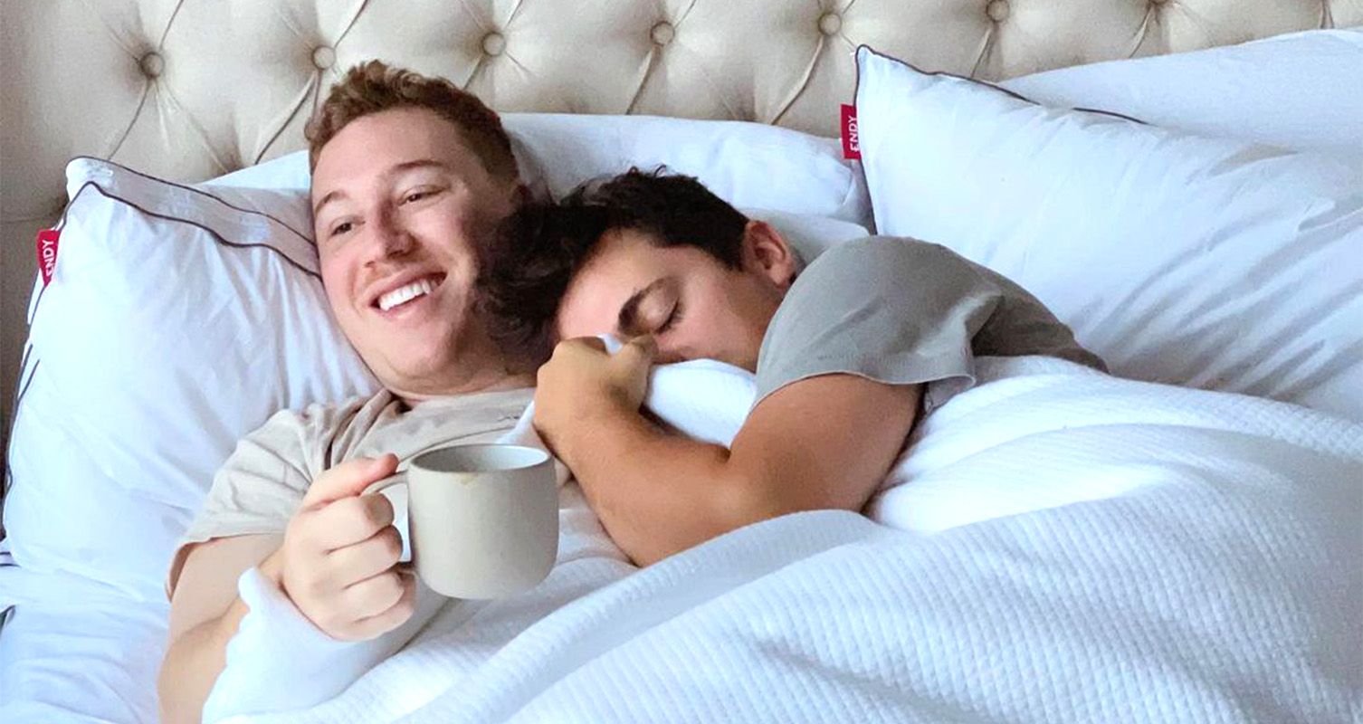 Two men cuddling in bed, one holding a cup of coffee.