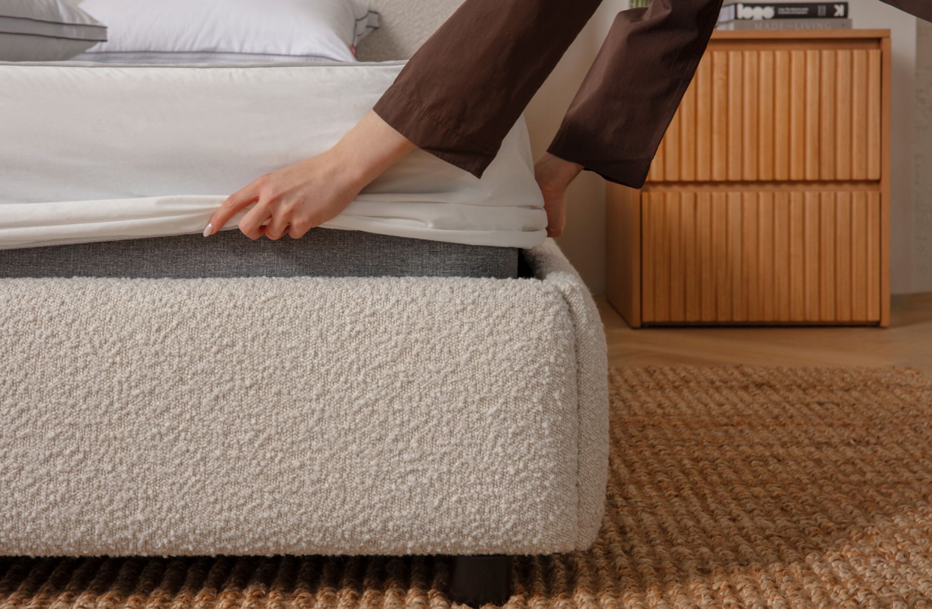 Close up of the corner of a bed, hands reach in from out of frame to put a mattress protector on a mattress