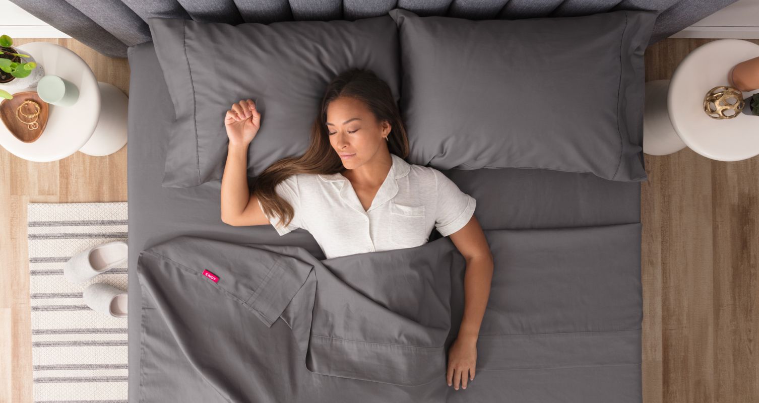 A woman sleeping in her bed, made with Endy Organic Cotton Sheets in Storm Grey, with one arm raised near her head and the other by her side.