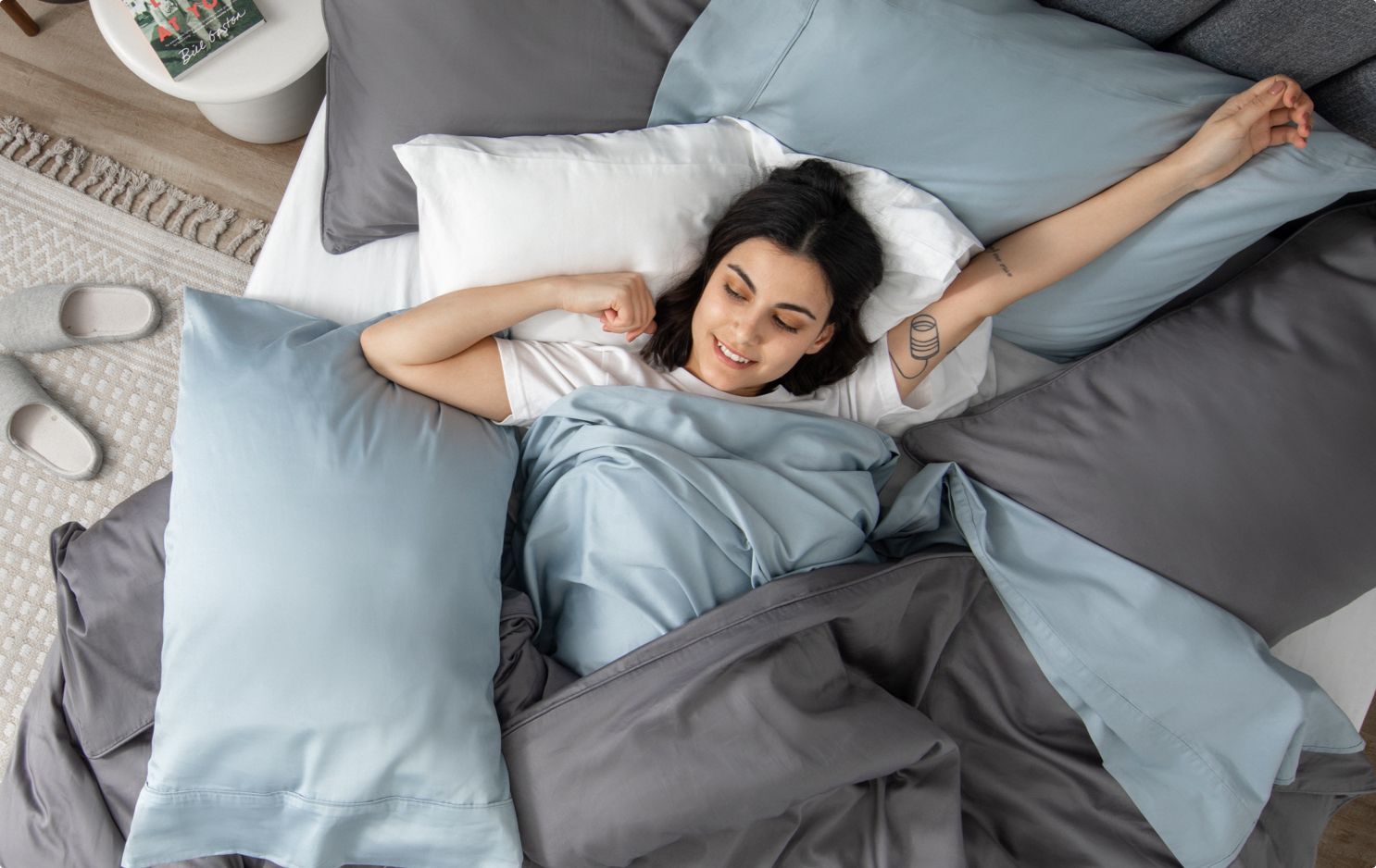 Woman stretching in a bed with blue sheets and pillows, and a grey duvet cover