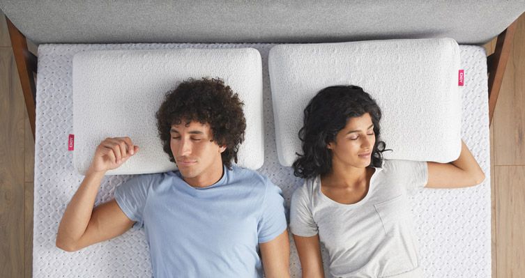 Two people resting on a bare mattress with two memory foam pillows on it.
