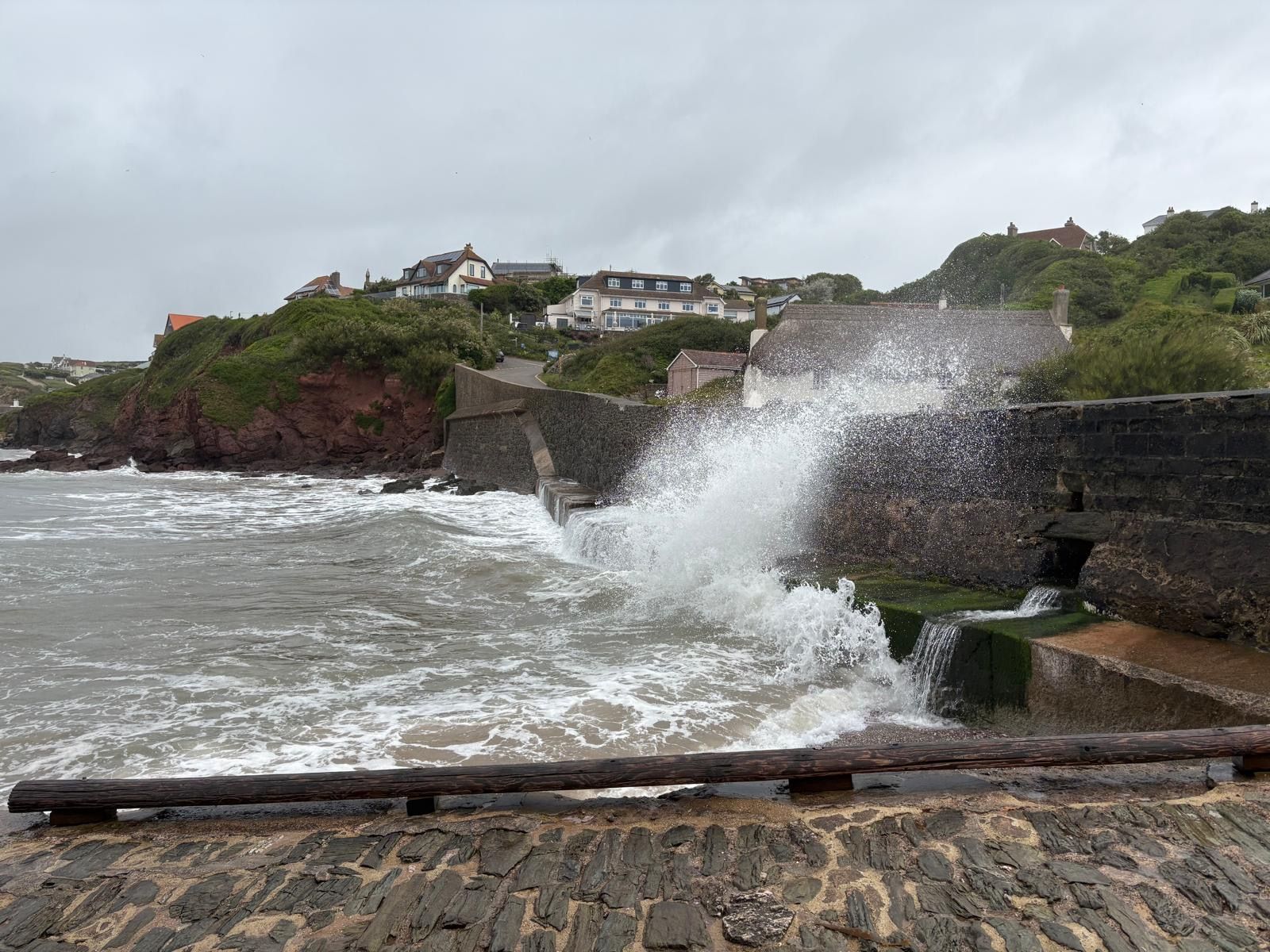 Stormy seas crash against the sea wall at Bude, Cornwall, with waves breaking dramatically along the rugged coastline and coastal homes overlooking the shore.