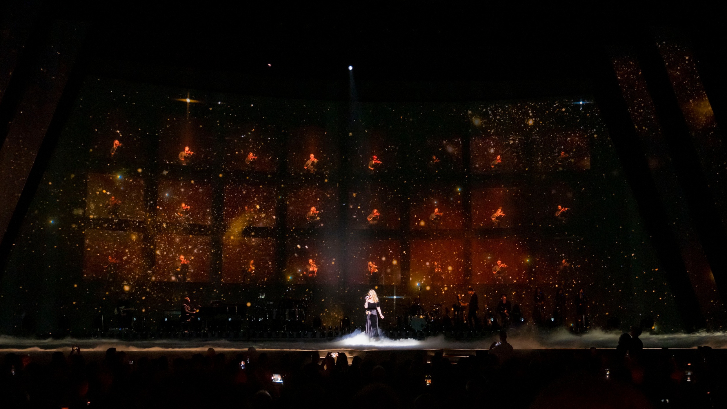 Singer Adele performing on large stage with band against a sparkling backdrop and orchestra on screen
