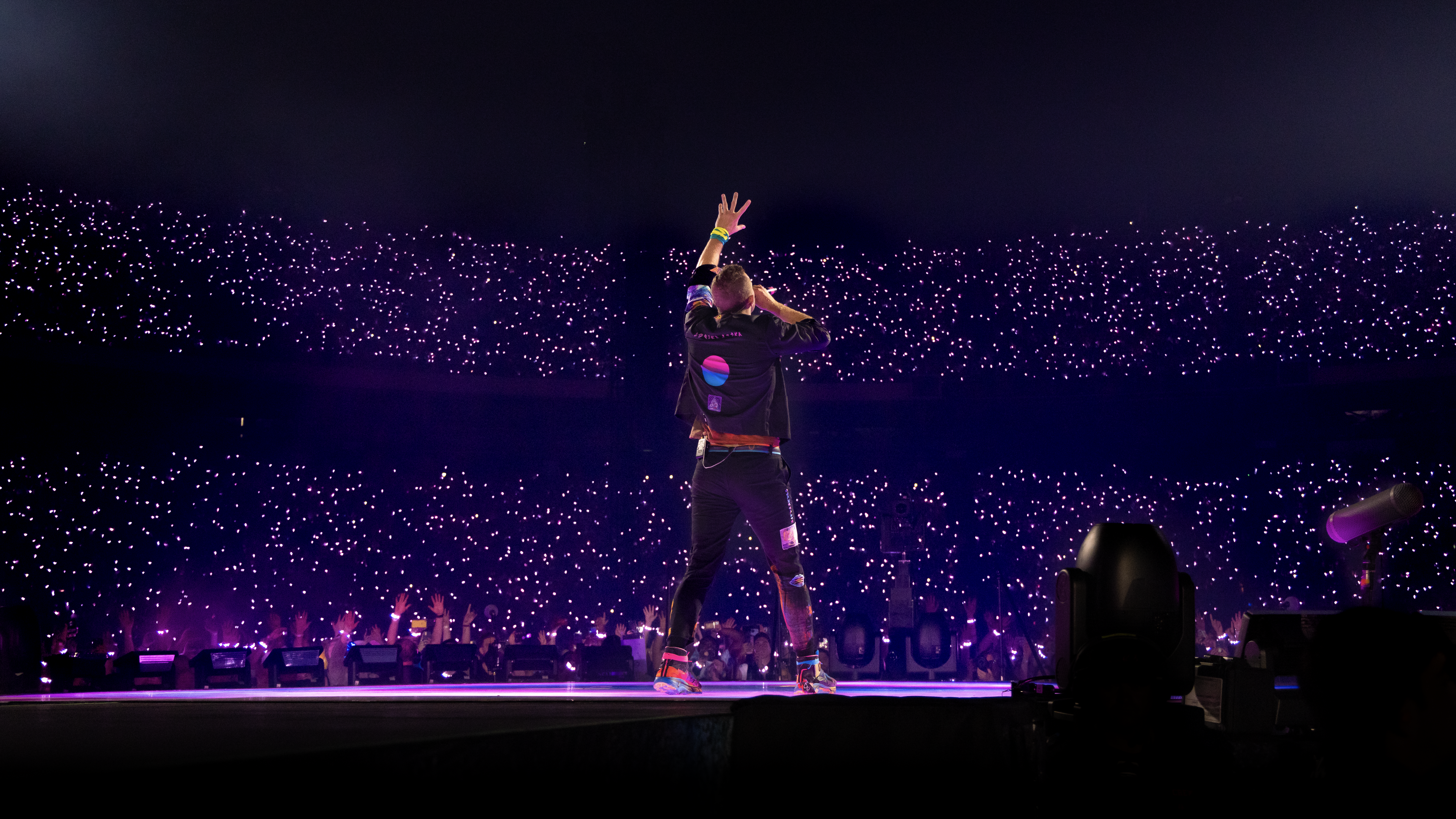 Back of Chris Martin with hand raised singing in front of lit up stadium crowd