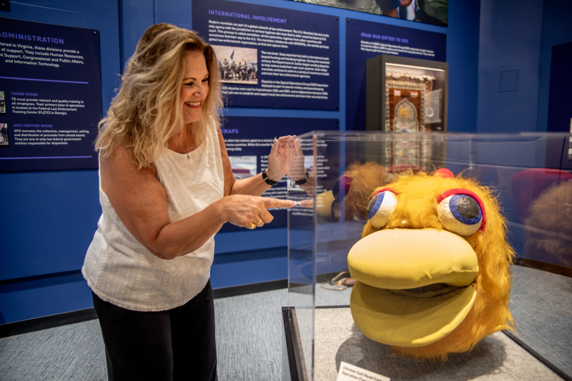 A museum-goer smiling and pointing at a podium with a glass box displaying a large textile head of what appears to be a yellow duck with cartoony eyes