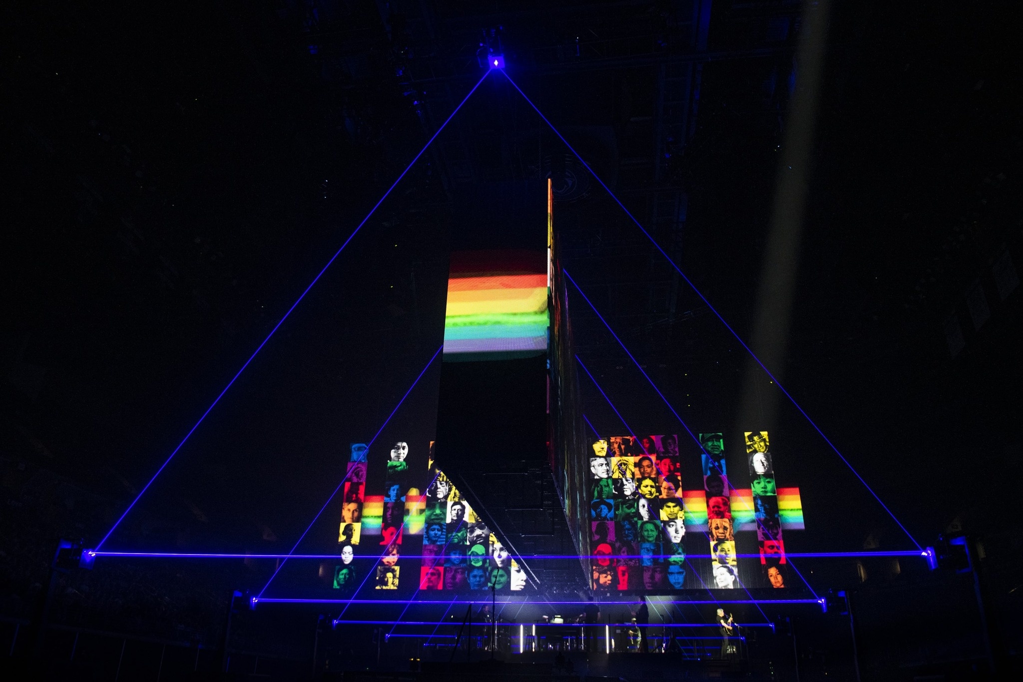 side view of stage graphics at the Roger Waters This Is Not A Drill Tour, graphics include blue triangles that seem to be floating in space, and a collage of multiple portraits of people in duotones, rainbow colors