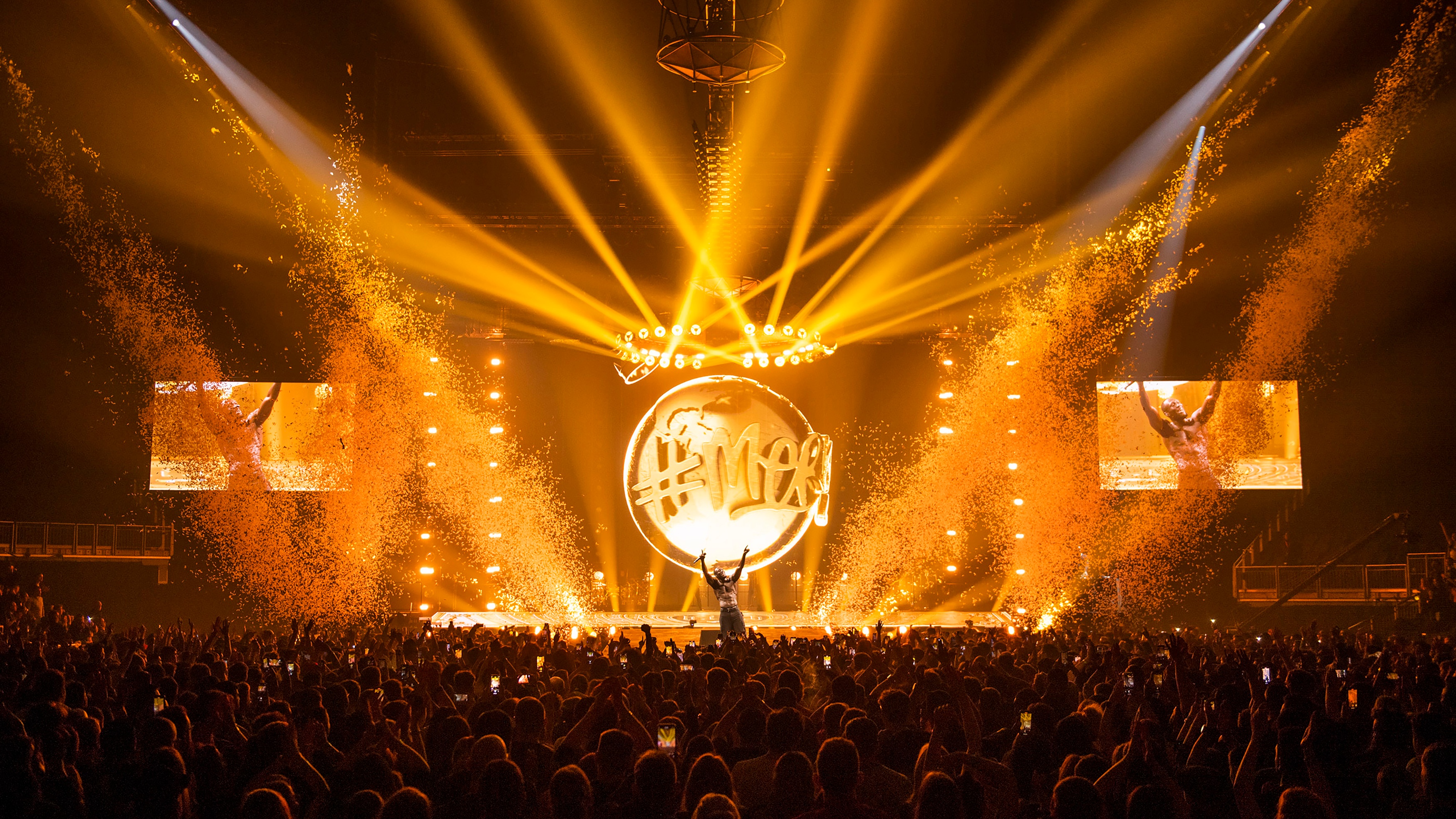 Rapper Stormzy performing on stage with gold lighting with screens on either side of himself and cheering crowd