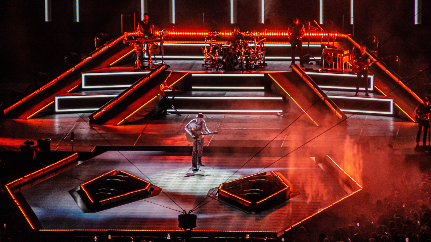 close up of performer playing guitar with the rest of the band behind him on a tired structure outlined by red and white lights