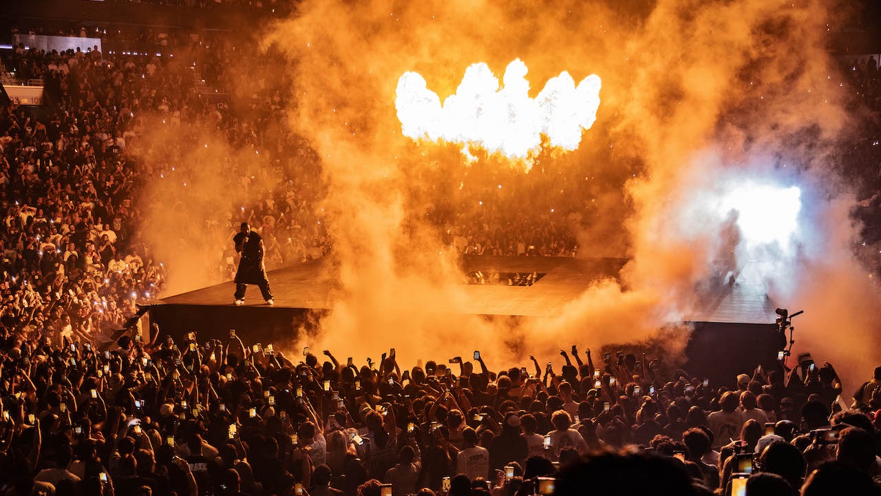 Rapper Drake performer on edge of stage in front of crowd with fire and smoke behind him