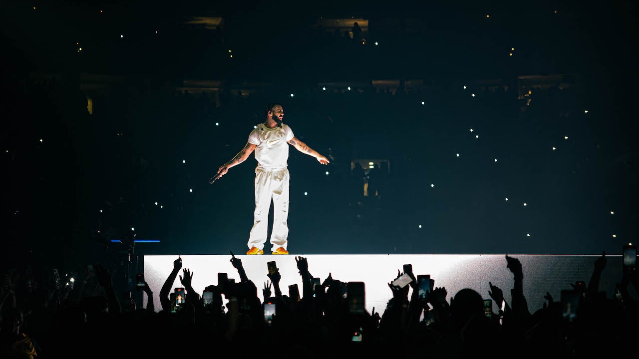 Rapper Drake in white jump suit standing on stage with arms and crowd reaching up