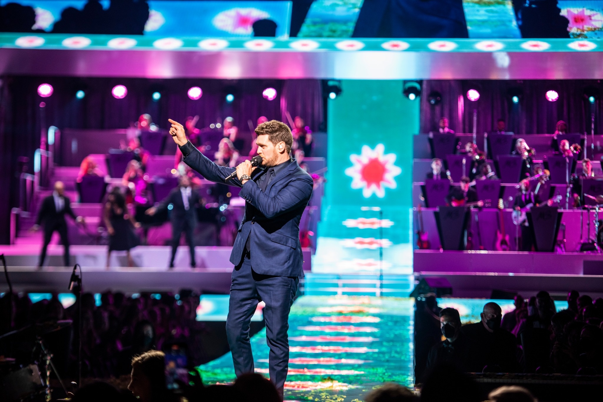 Close up of Michael Bublé performing on stage, looking to the left, one arm raised, finger pointing up, during the The Higher Tour, in the background the orchestra is visible, lit up in purple