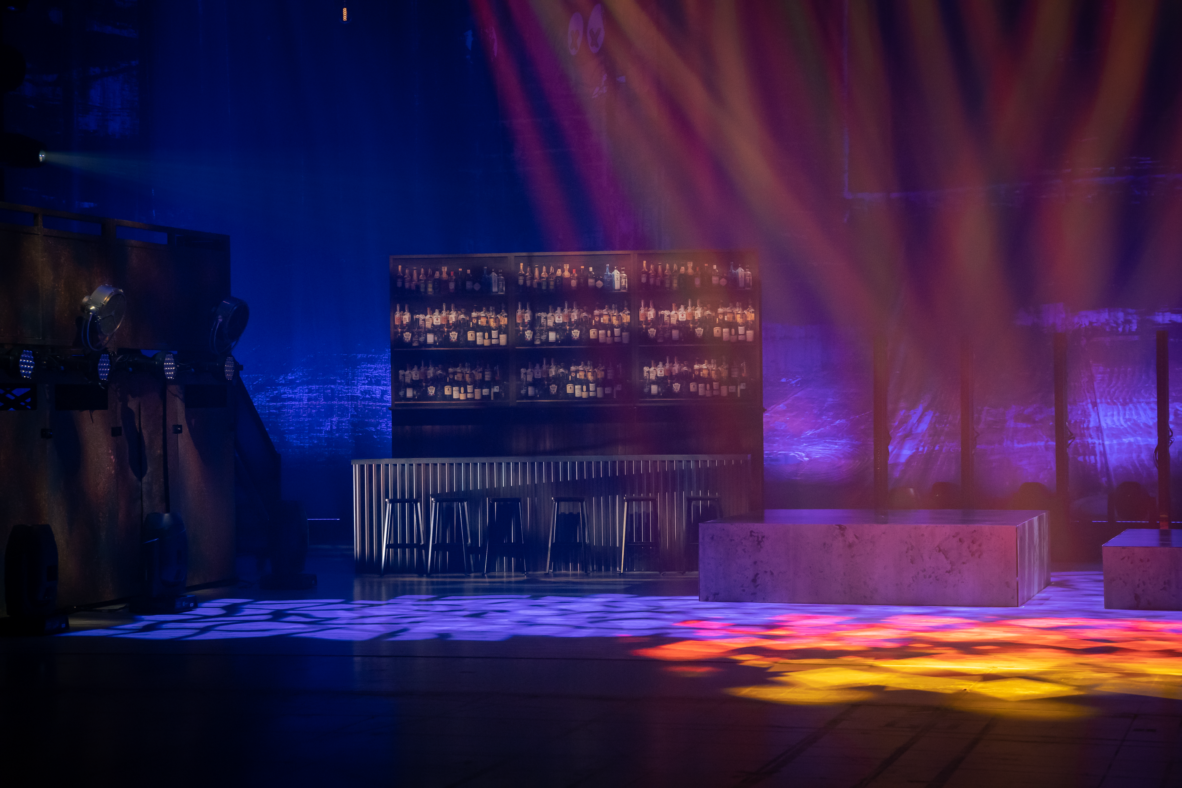 Black Crowes Shake Your Money Maker Tour stage design featuring a brick wall backdrop, a standalone bar with stools and alcoholic bottles in the shelves, stage is light up in blue lights with pink and yellow