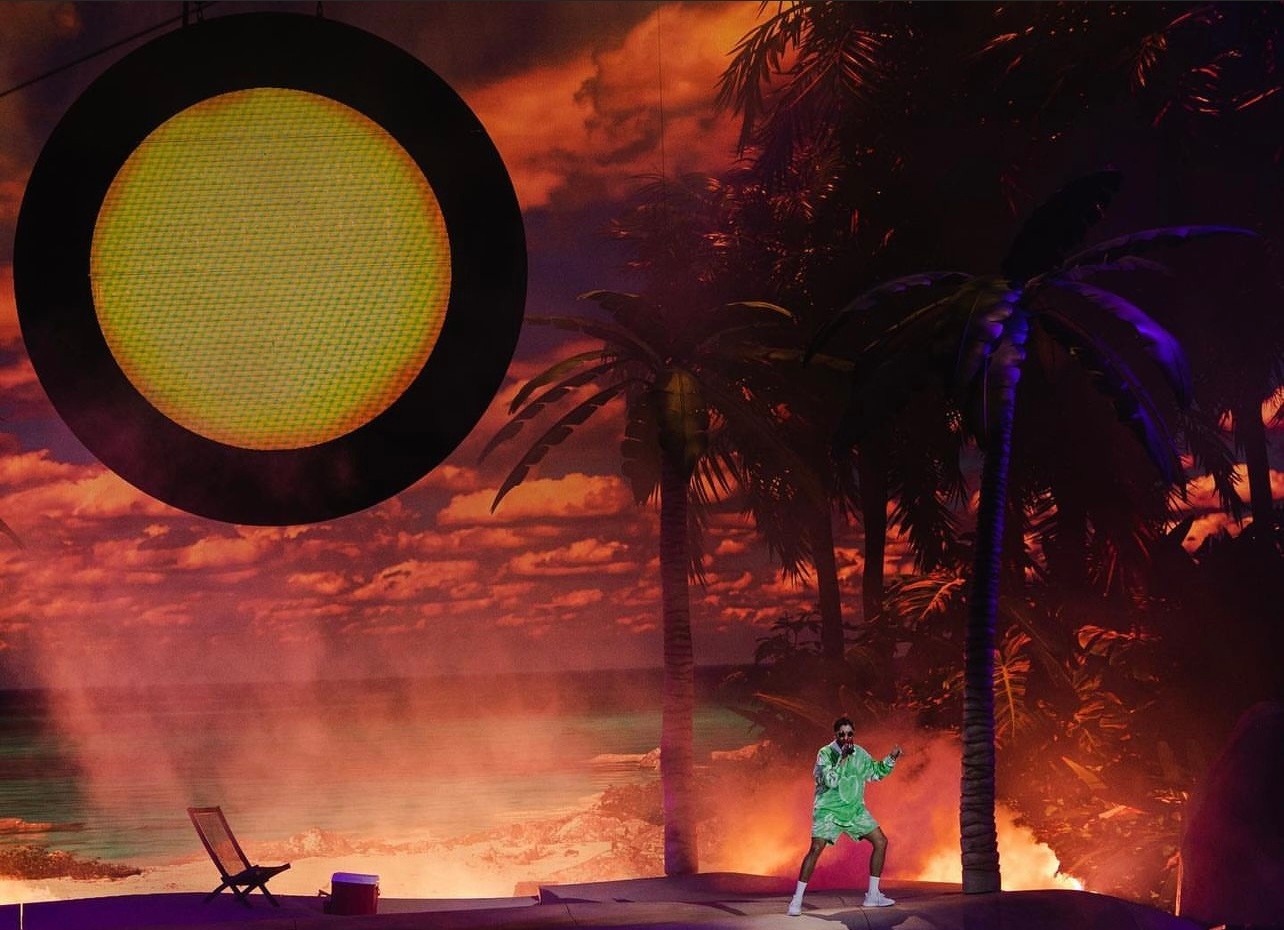 Bad Bunny performing on stage with palm tree and beach setup behind him
