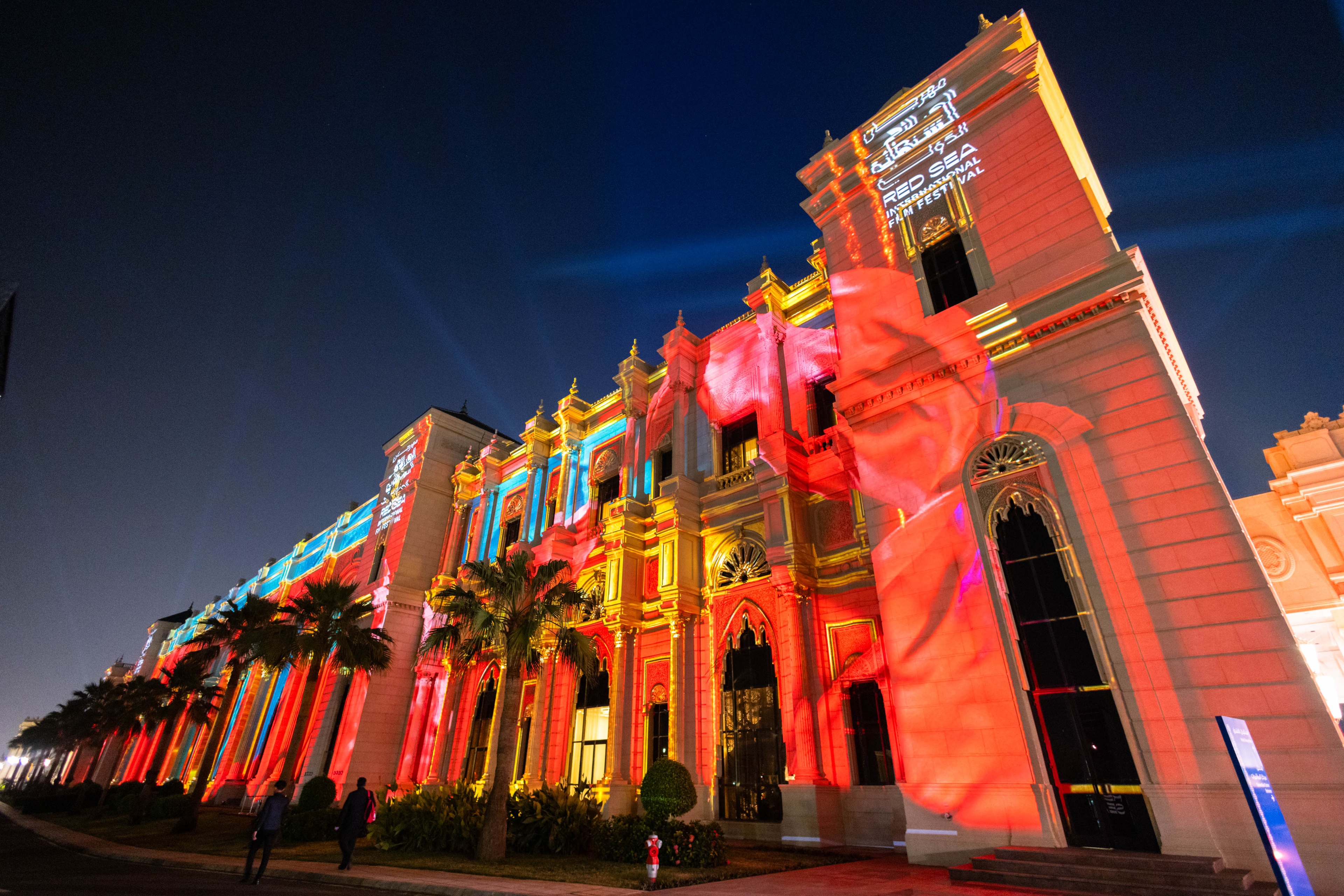 Beige building lit up peach and blue with palm trees out front