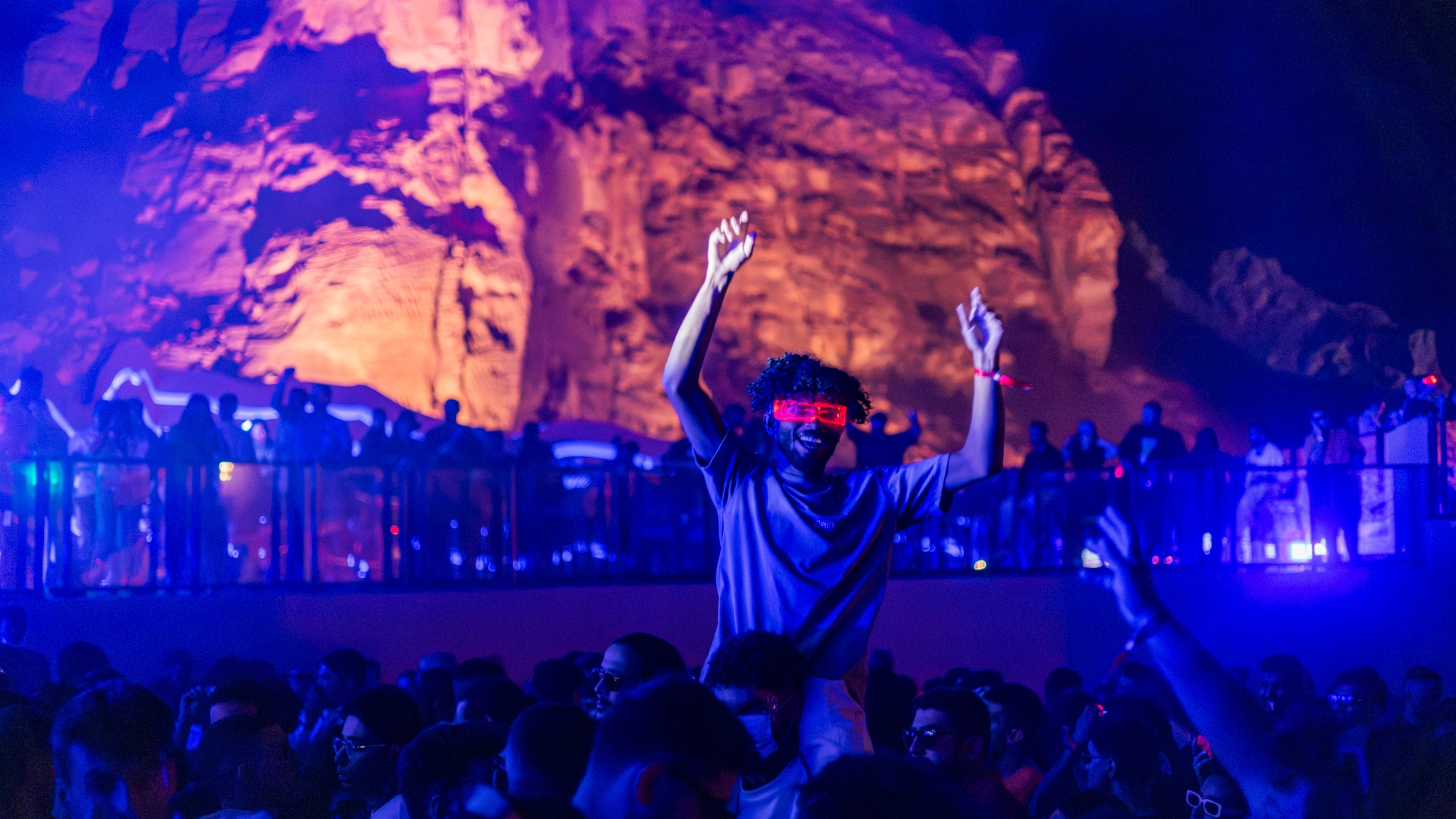 Person wearing fluorescent eye mask above crowd in festival in rock setting low lighting