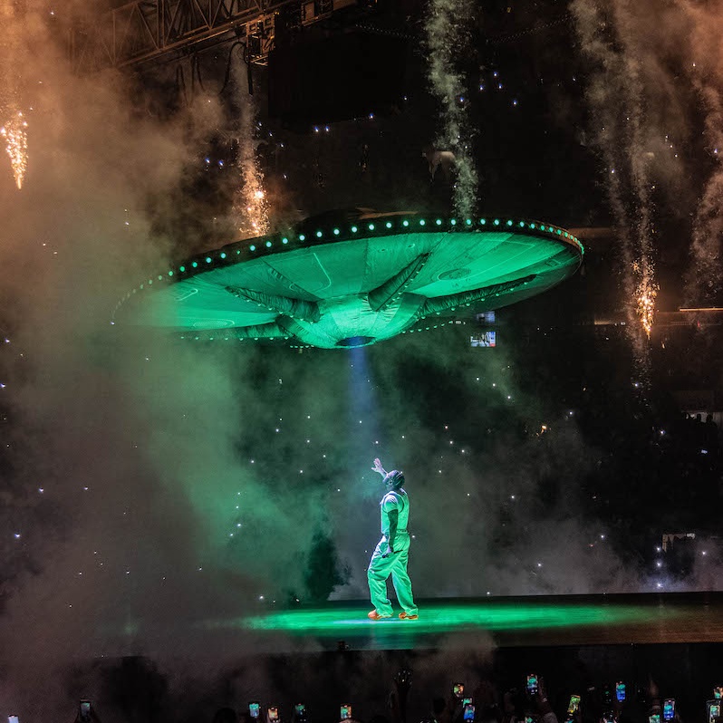 Rapper Drake performer on stage lit with green lighting looking up at green space ship with crowd