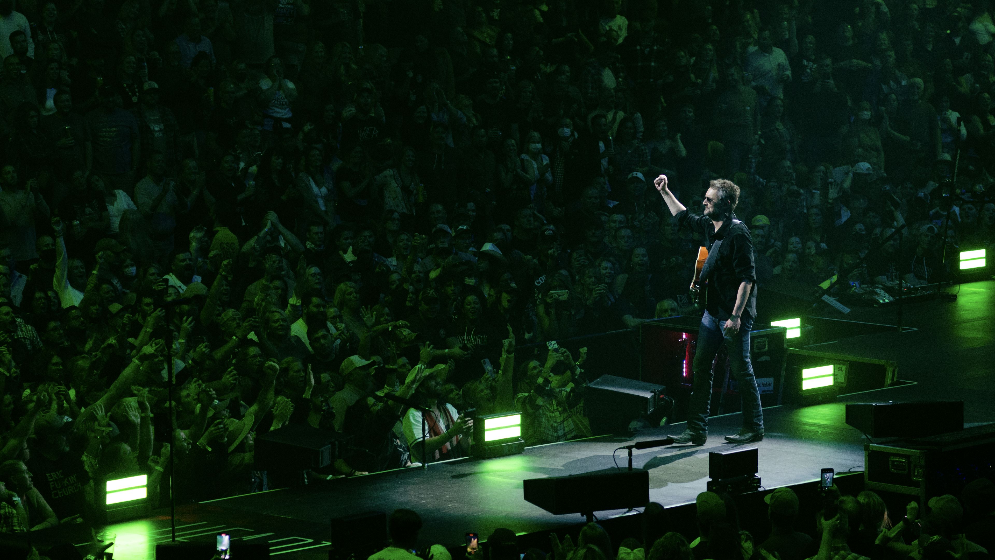 Eric Church walking on stage with his guitar on the back and one arm up fist closed in the air signaling victory, crowd around him cheering with arms up in the sky