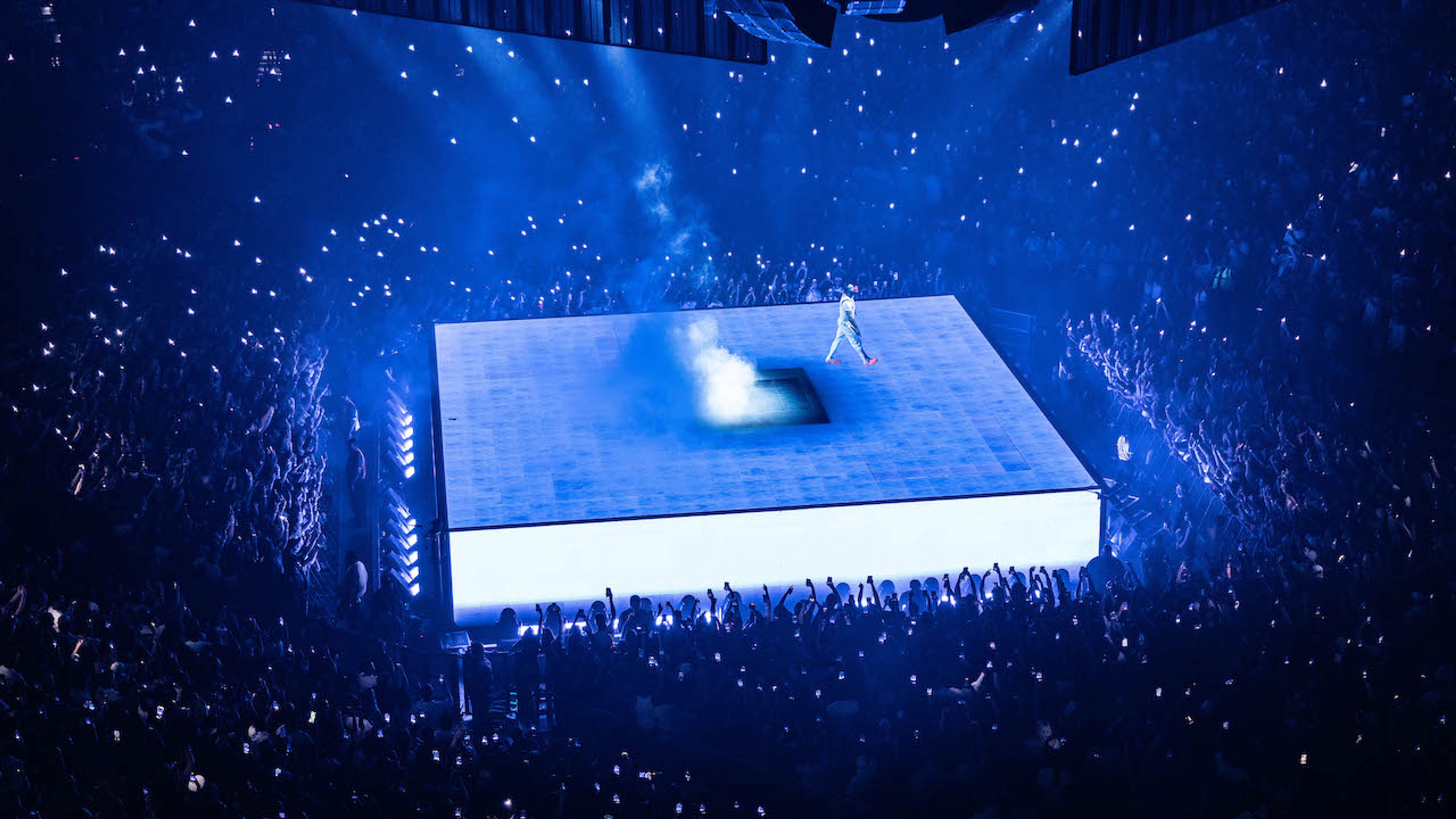 Rapper Drake performer on larger LED screen square platform with blue lighting surrounded by spectators