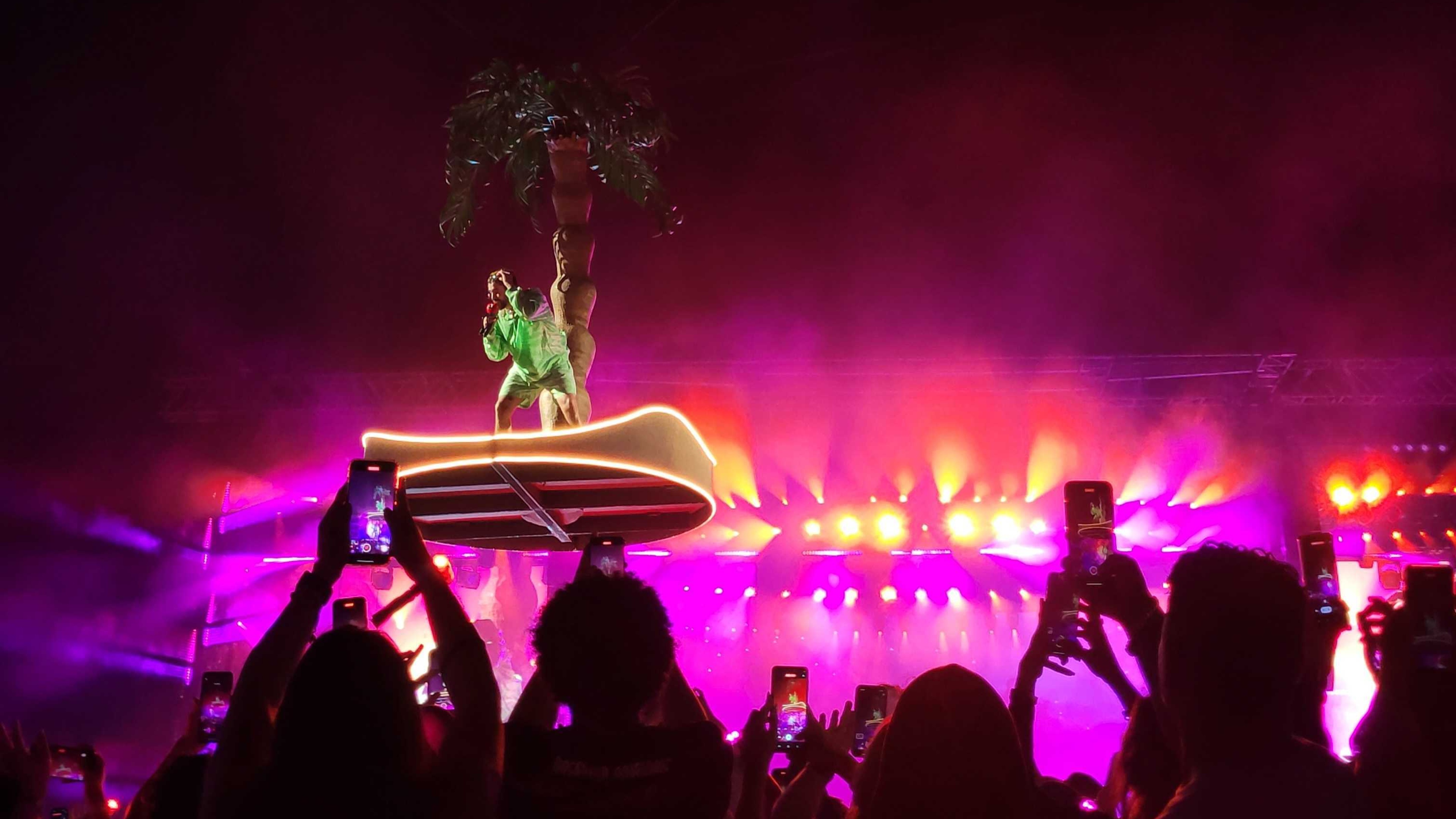 Bad Bunny flying above cheering crowd on palm tree with pink lights behind him