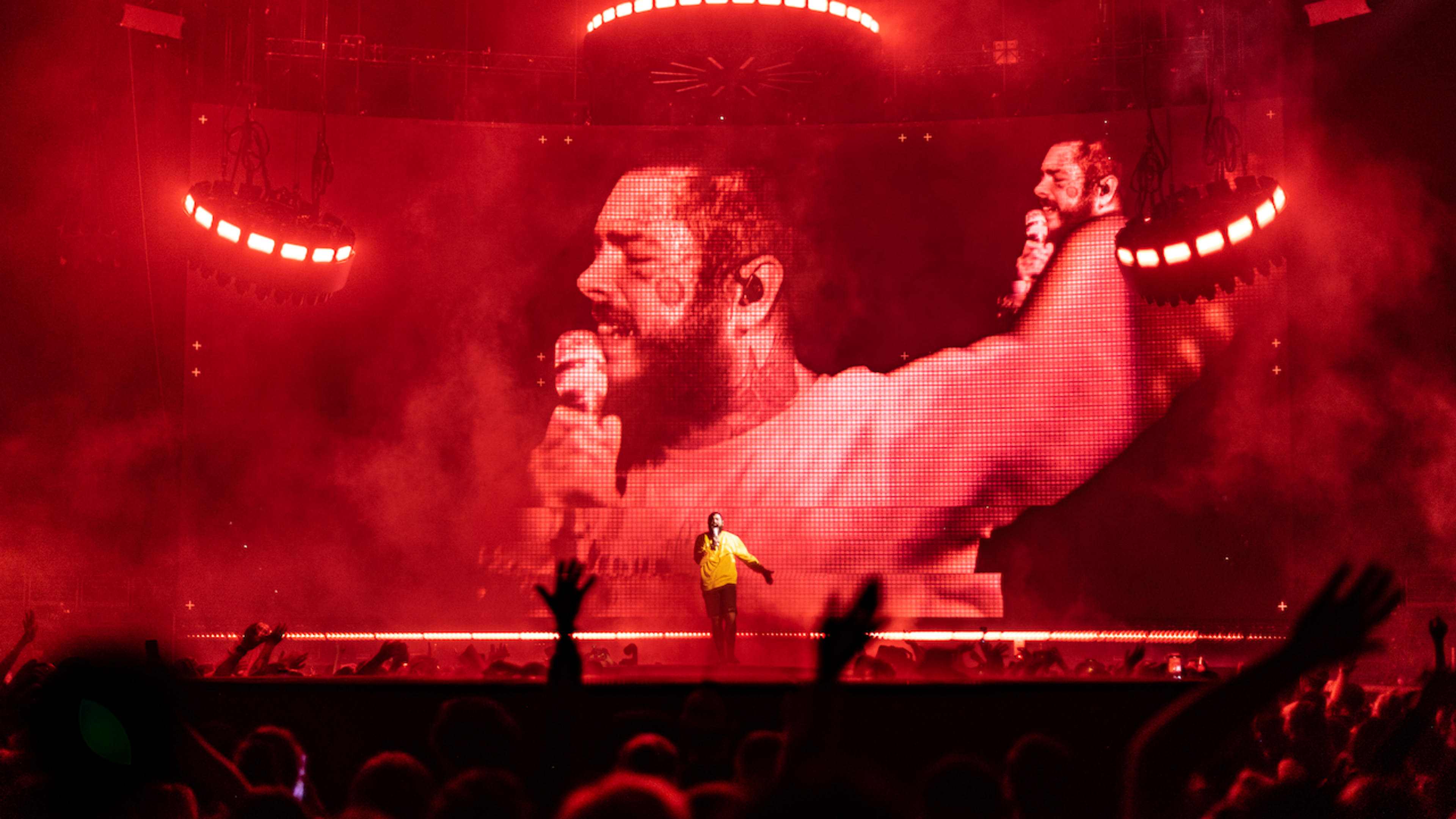 Post Malone performing on stage with yellow shirt with large LED screen lit up red behind him