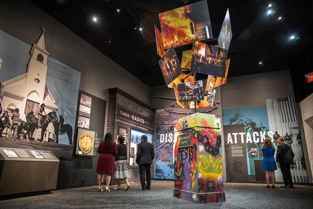 Museum experience of historic replica building facades, photos, text, and in the foreground a large sculpture of a public statue bottom with graffiti with a growing stack of protest photos resembling a fire