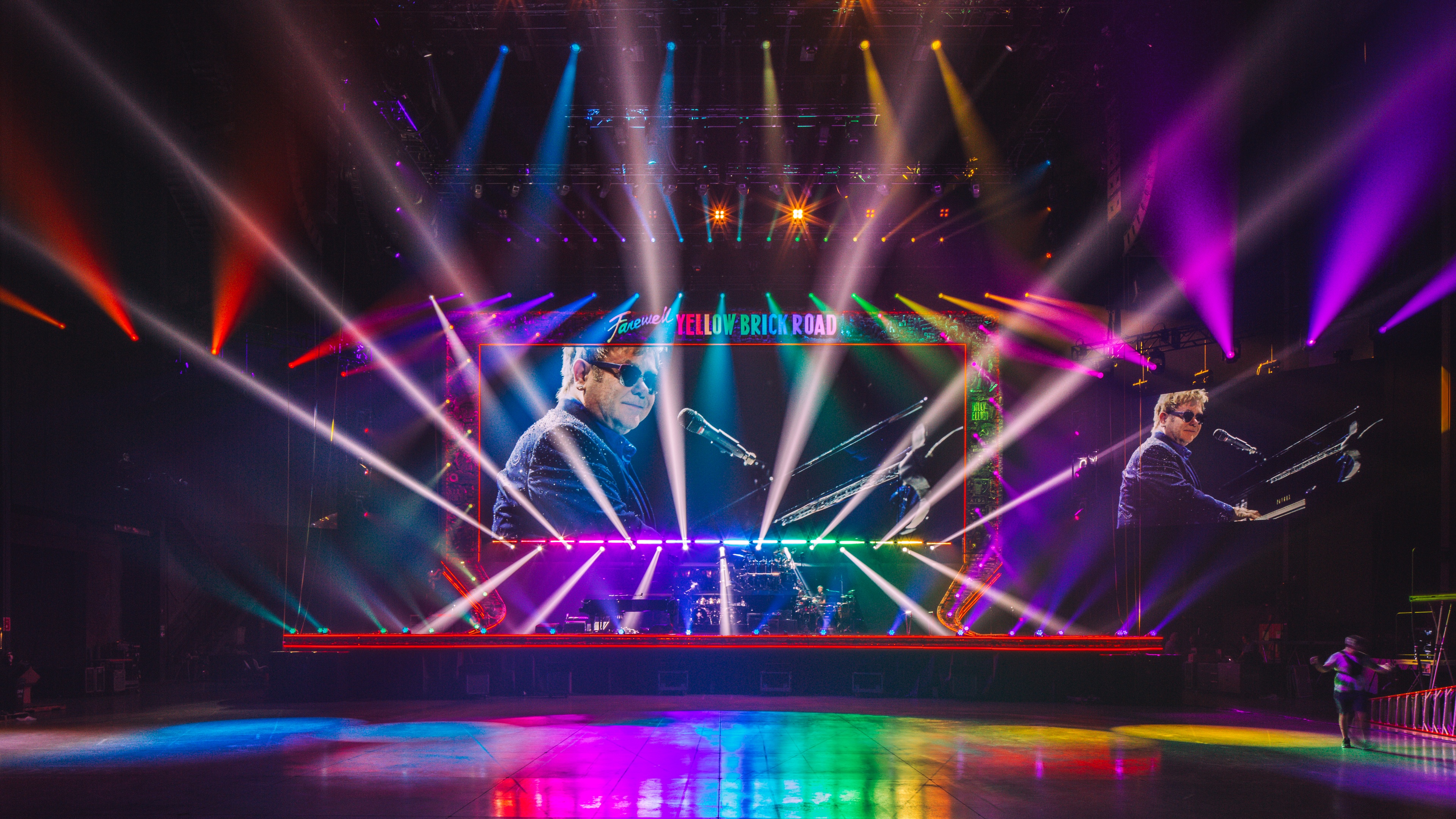 Elton John on stage performing on piano with rainbow colored lights