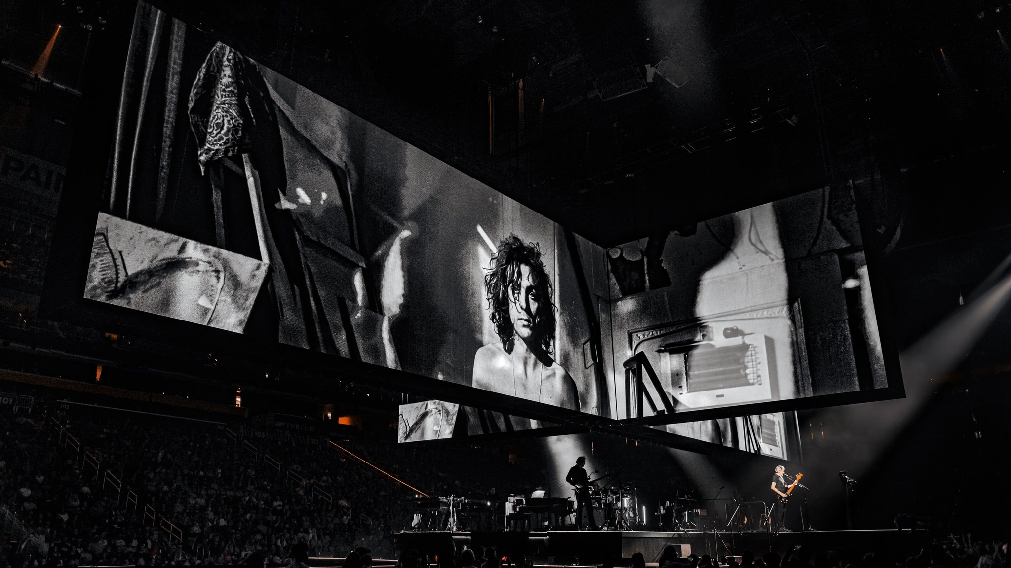 crowd watching Roger Waters and his band play on stage during the This Is Not A Drill Tour, big screens above the performers feature a black and white photograph of a young roger waters