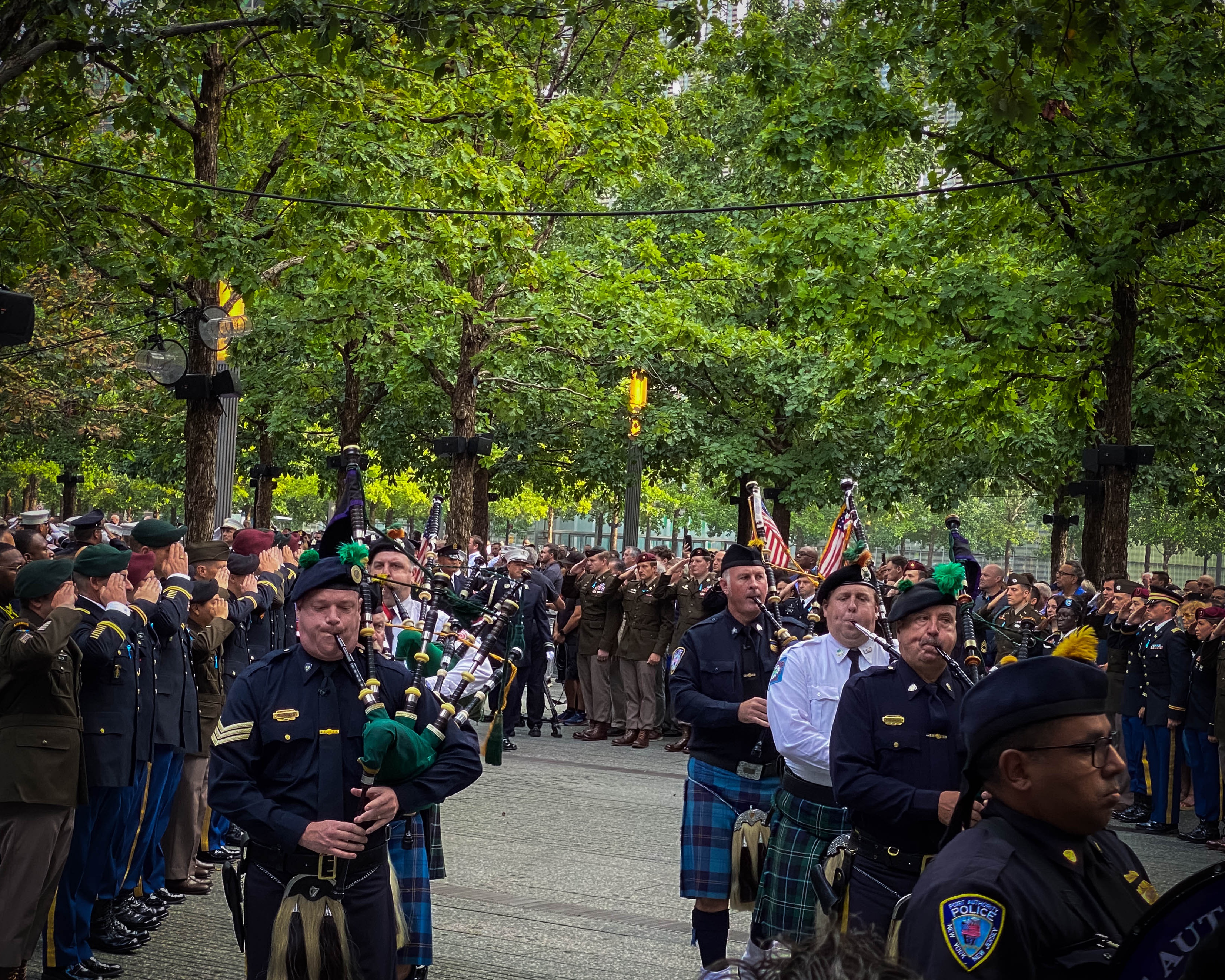 Military figures walking through park playing bagpipes surrounded by saluting military people