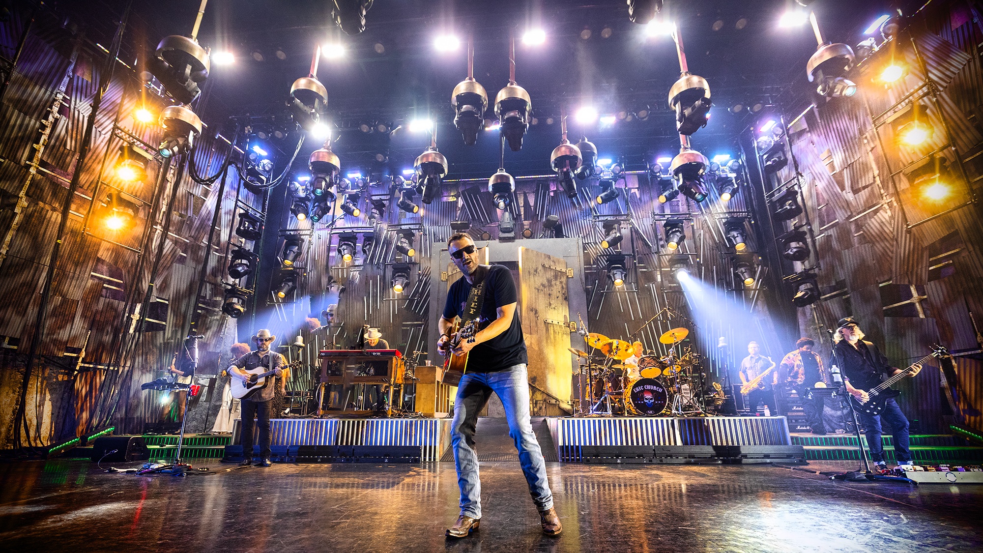 Front view shot of country artist Eric Church performing on stage with band, light show and hanging lights
