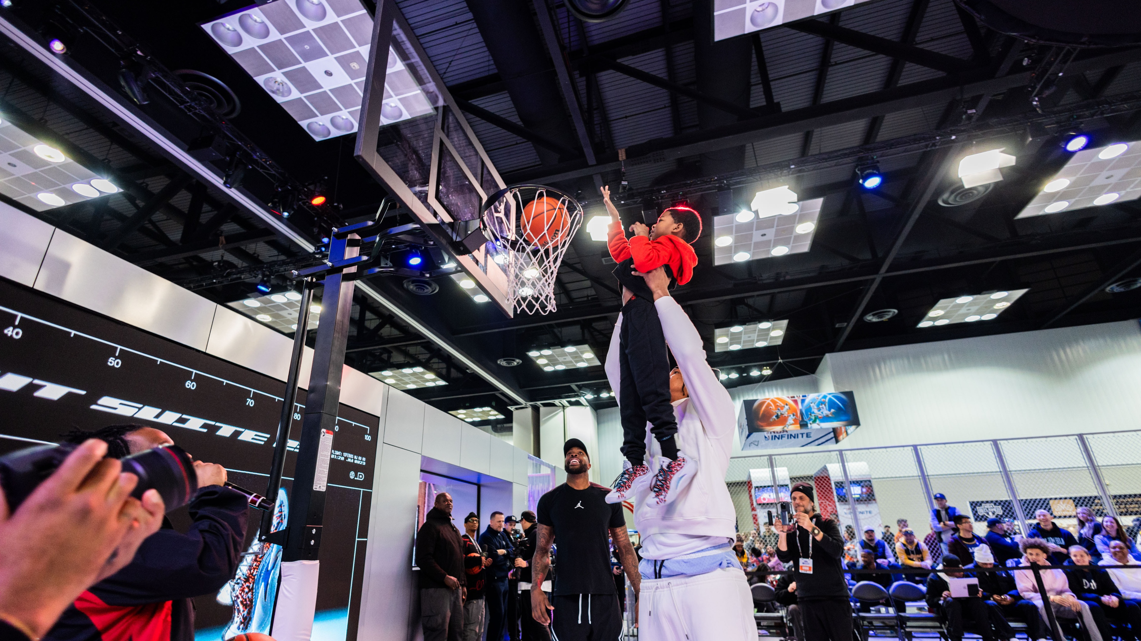 Basketball athlete holding child up to put the basketball through the net with crowd of people watching and photographers