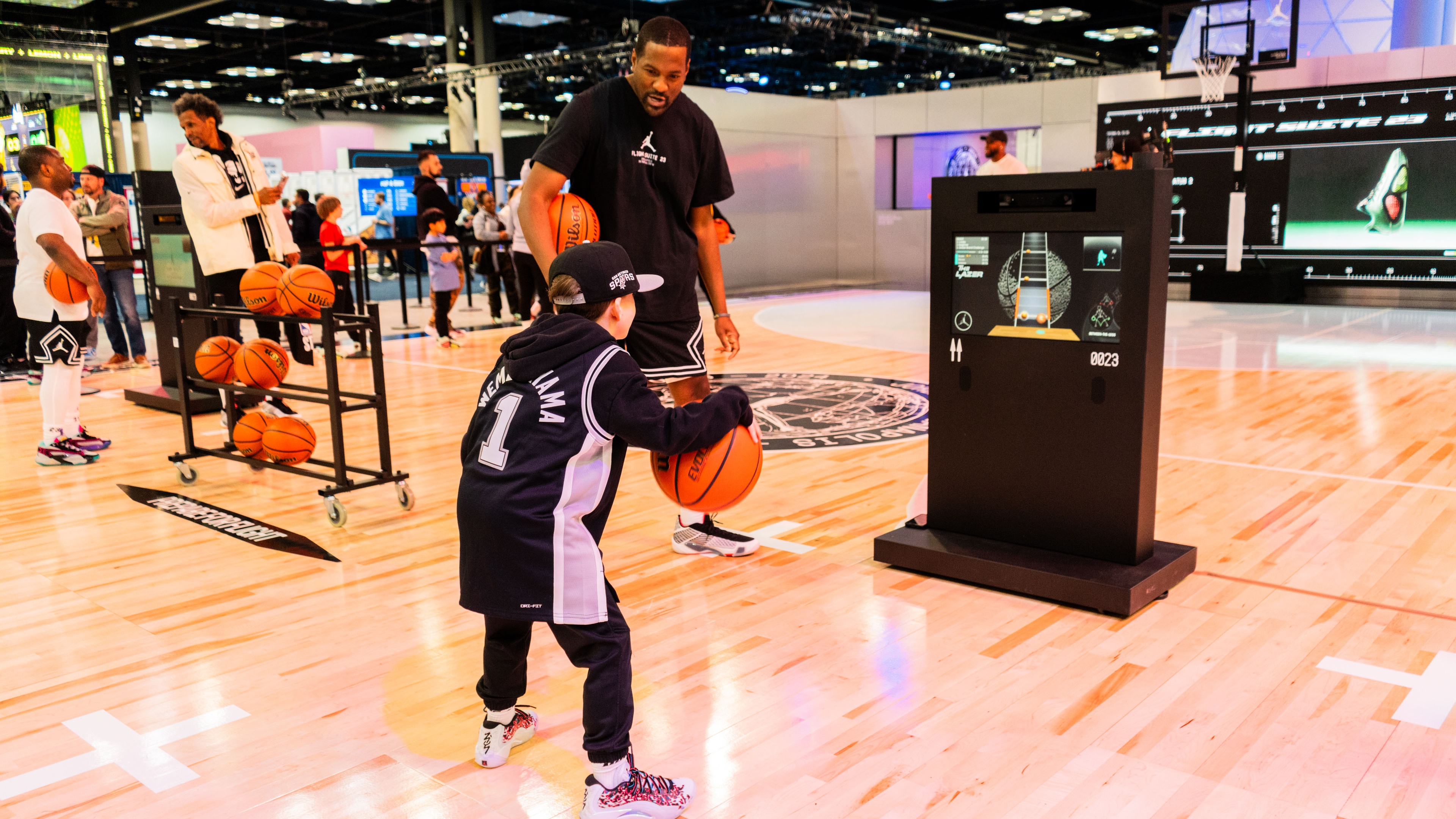 young person dribbling basketball infront of interactive game on screen on basketball court watched by athlete