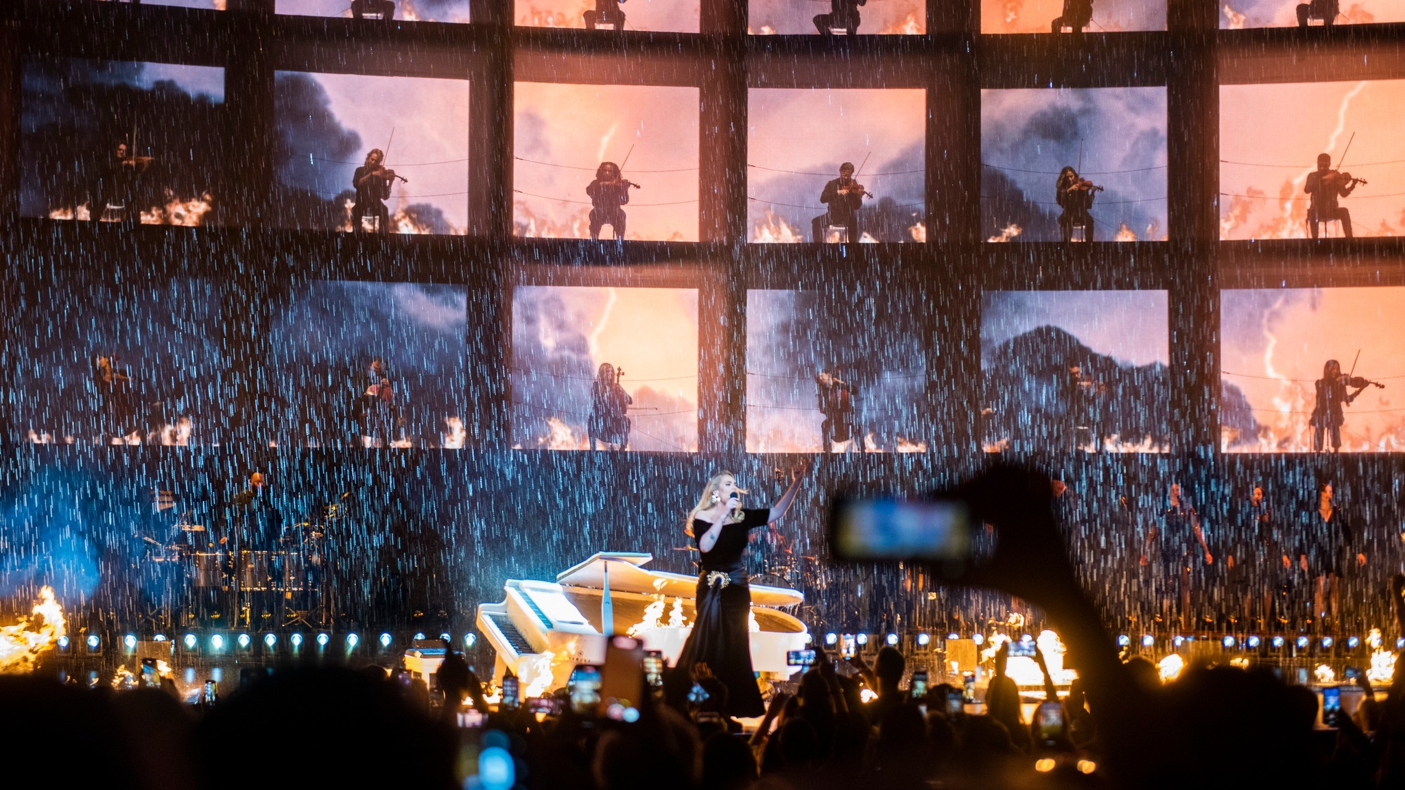 Singer Adele performing on stage in the rain in front of broken grand piano pyro effects and orchestra in a grid on LED screen