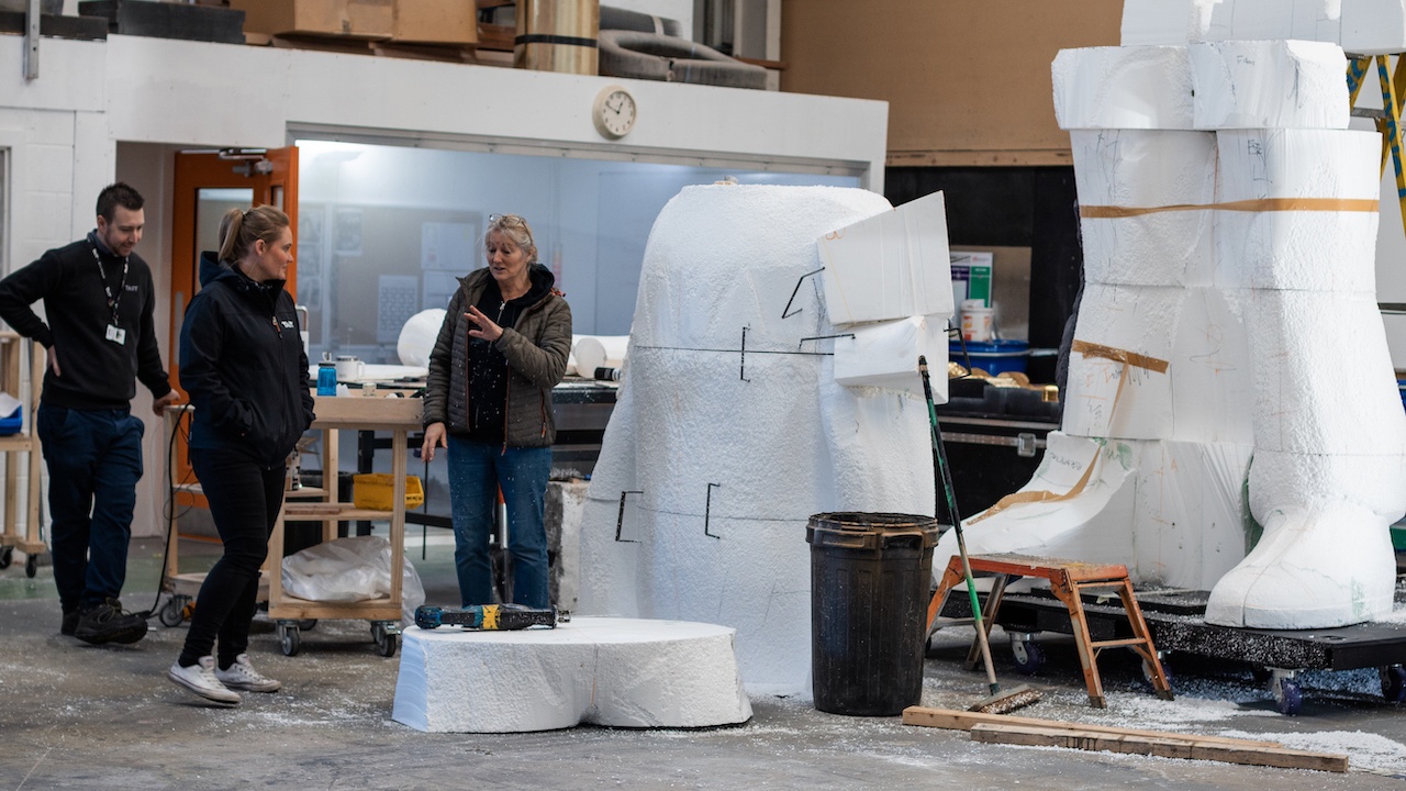Team of people in industrial setting talking next to parts of huge polystyrene sculpture