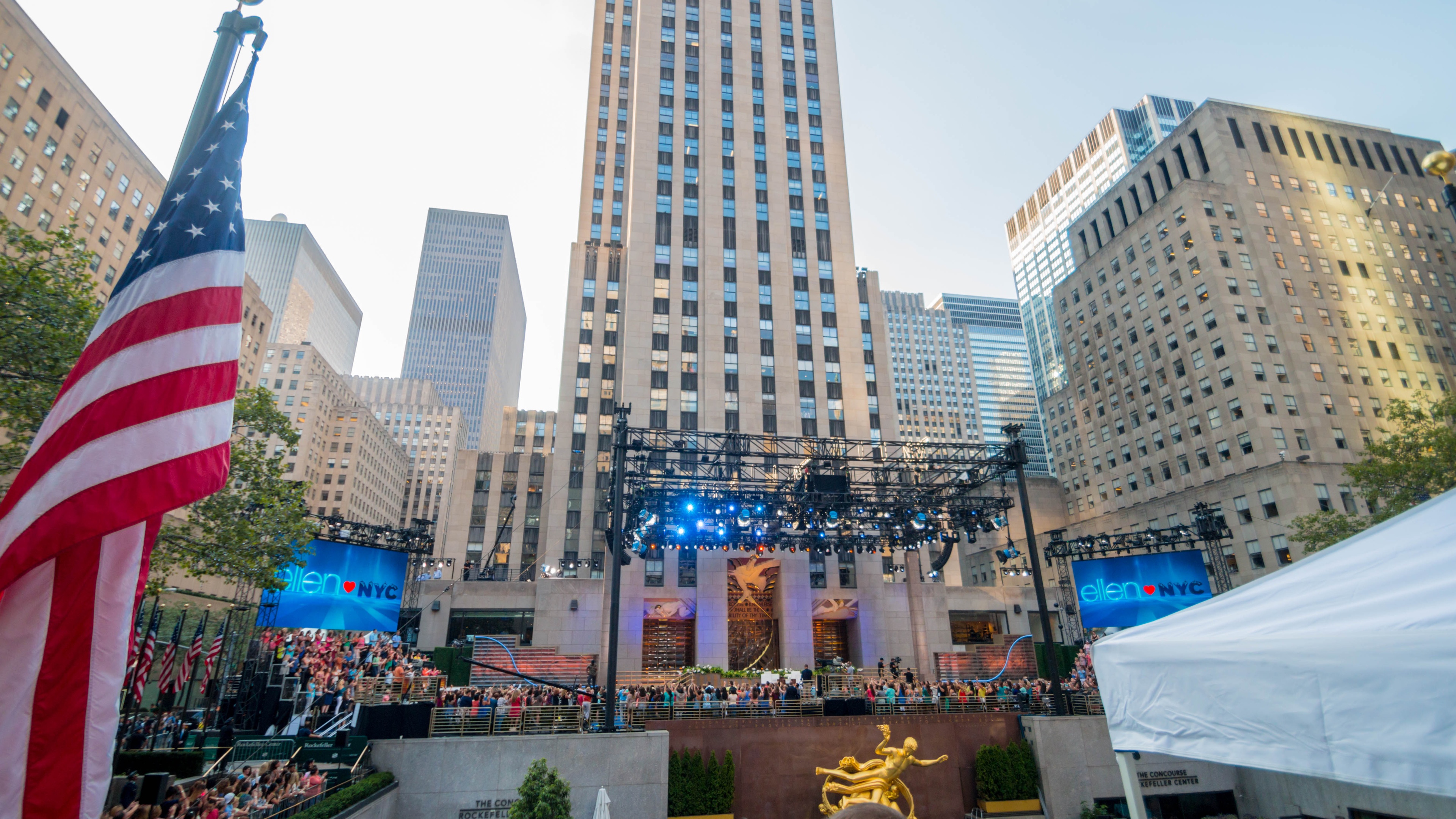 Rockefeller Center in the daytime, set up with a stage, blue lights, and screens and surrounded by an audience on risers