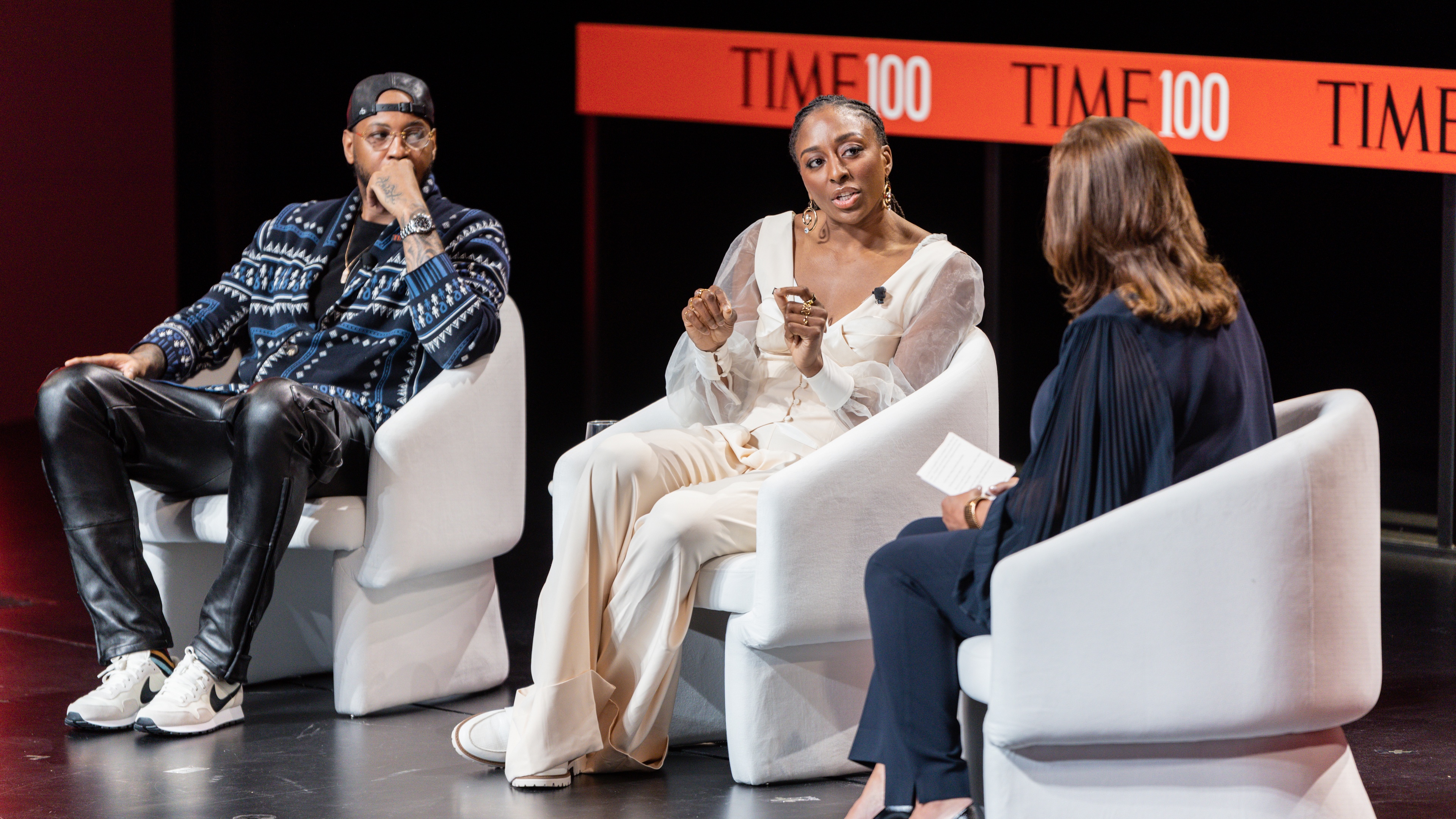 Three people on white chairs with TIME100 logo behind them on a screen