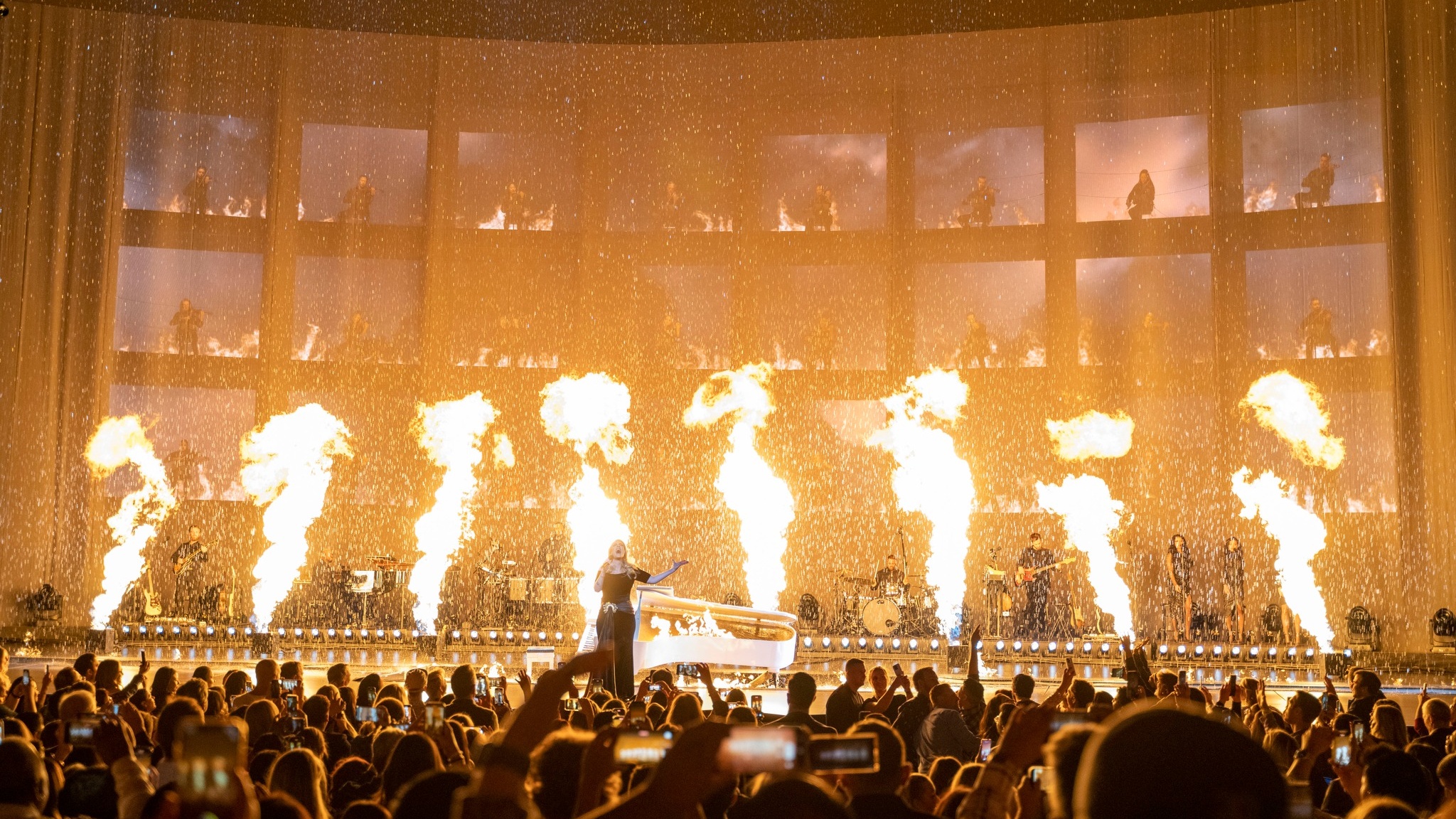 Singer Adele performing on stage in front of crowd behind her is a broken grand piano and pyro effects in front of full band and orchestra