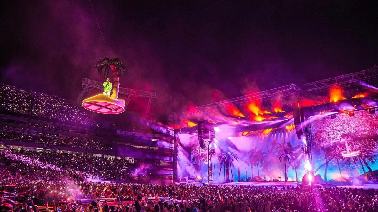 Bad Bunny flying above stage on palm tree lit up pink in blue with large arena crowd