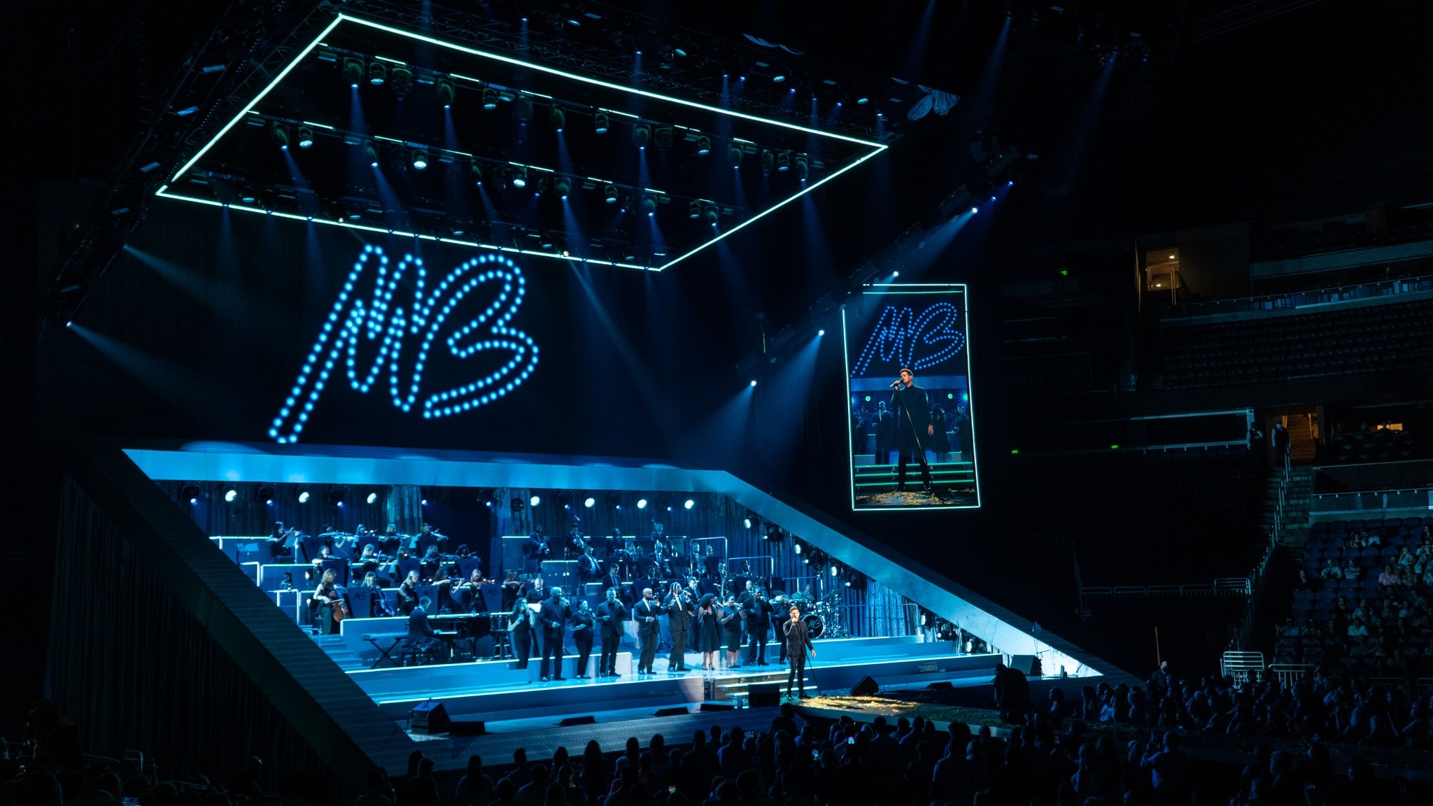 Michael Bublé performing on stage surrounded by backup singers and a live orchestra during the The Higher Tour stage, blue lights, screen to the rights shows a close up of michael Bublé, and above the stage in dotted letters shines an MB
