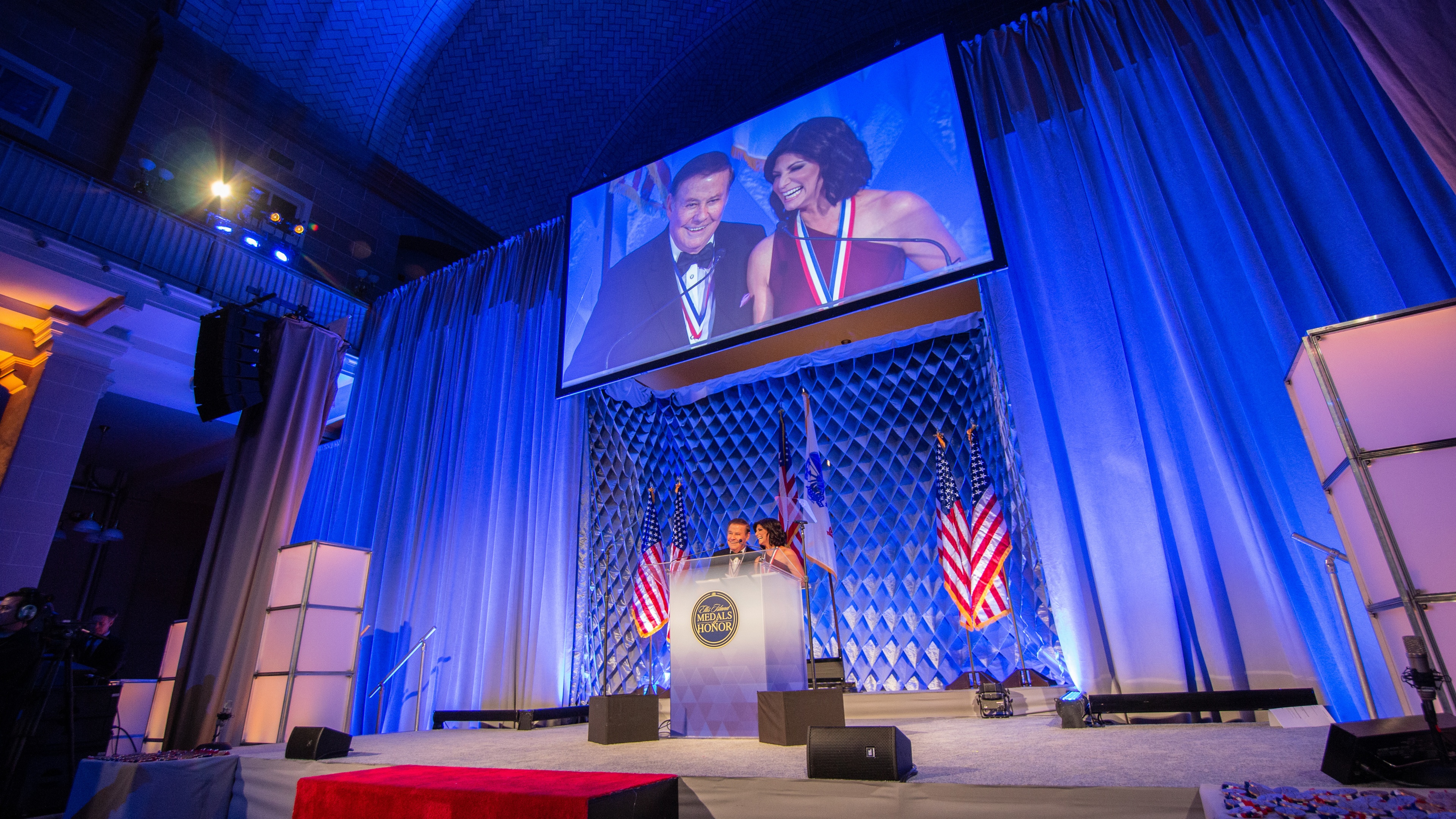 2 speakers wearing medals stood at podium with american flags in front of large blue drapes and LED screen