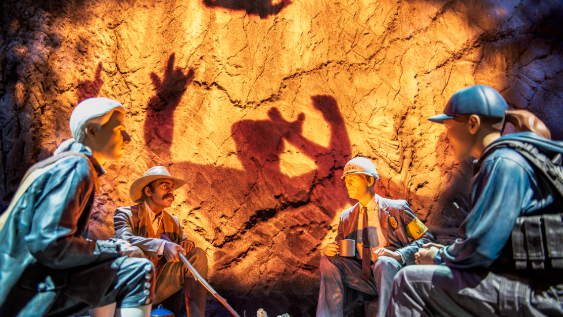 Mannequins sitting around a fake fire, shadows behind them on rock wall