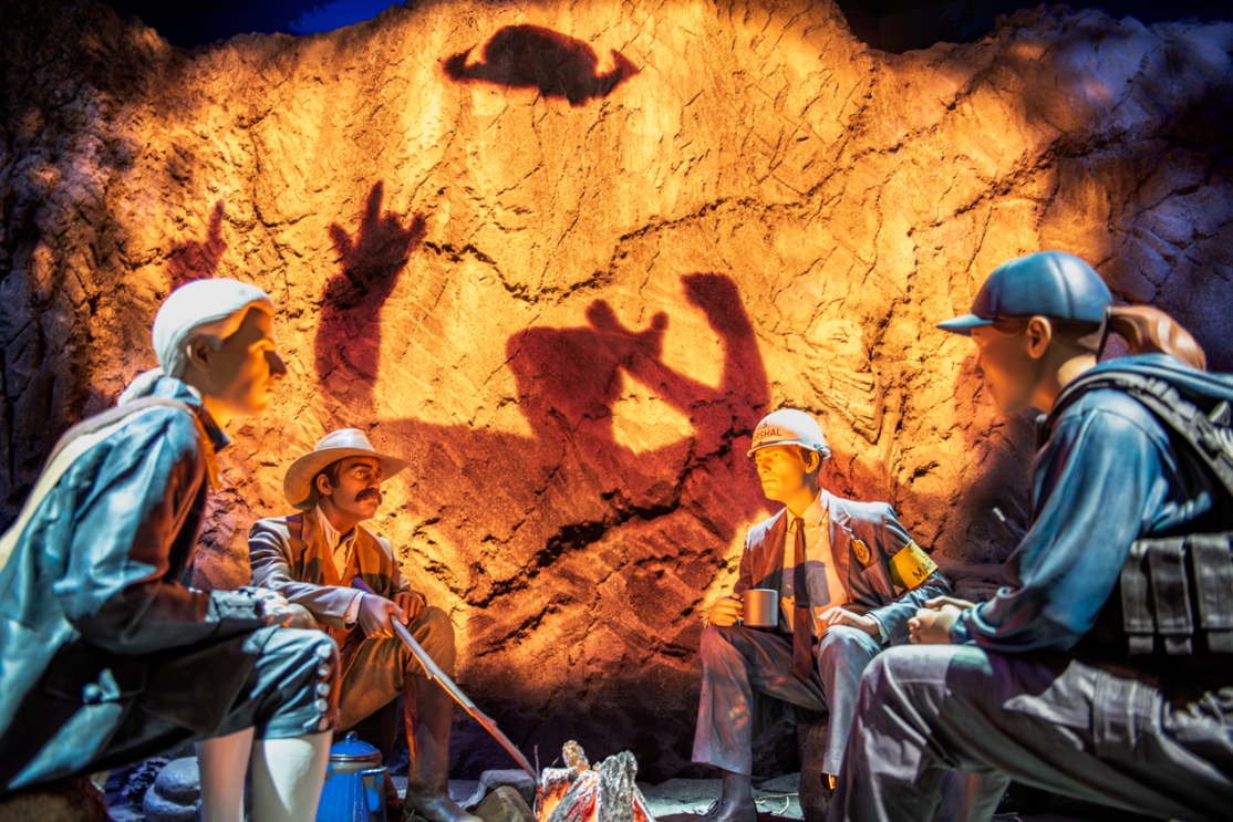 Museum display of realistic people statues sitting around a campfire with a silhouette shadow glowing on the rockface behind them