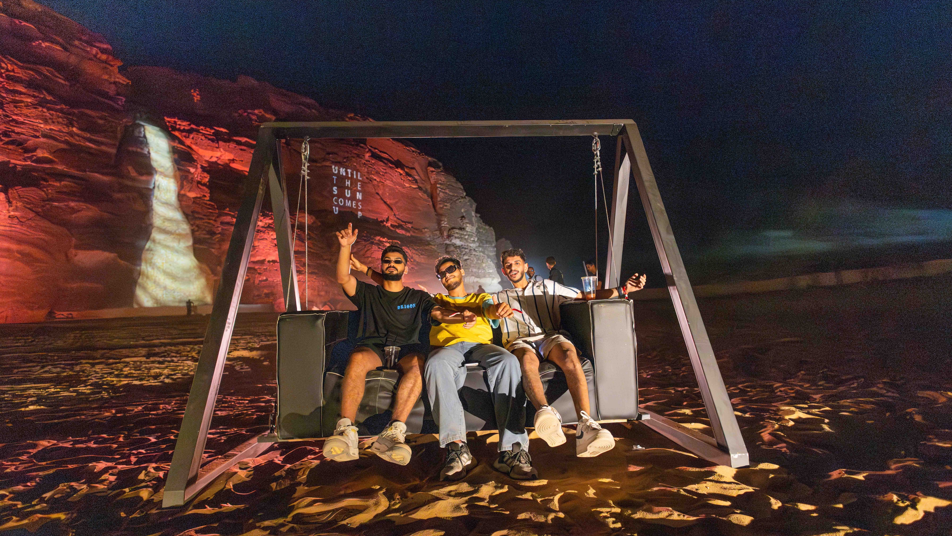 3 men on a wooden custom swing in the middle of the dessert at festival in front of rocky setting